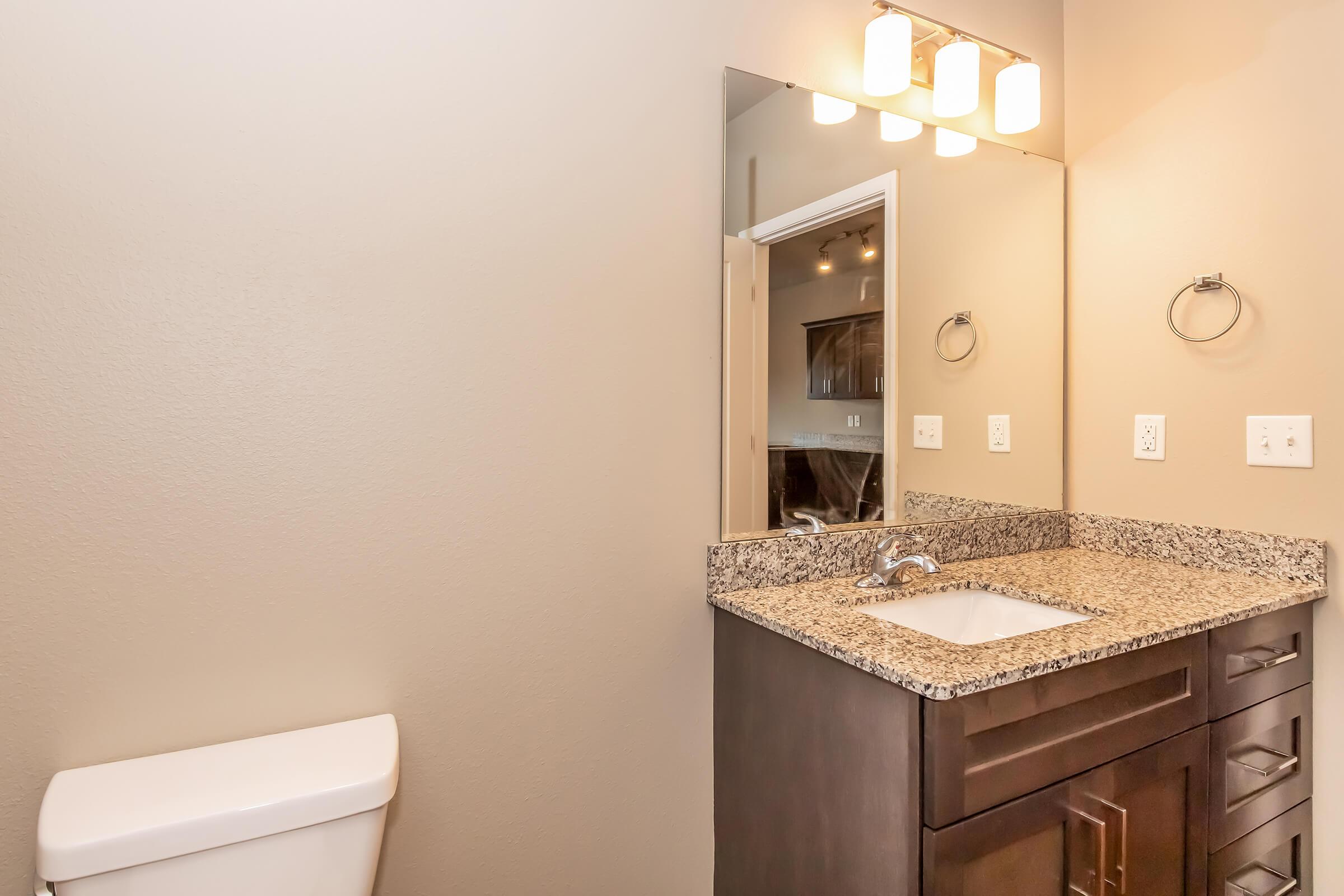 A modern bathroom featuring a granite countertop with a sink, a large mirror with four light fixtures above, and a towel ring. The space includes a white toilet and neutral-colored walls, creating a clean and contemporary look.