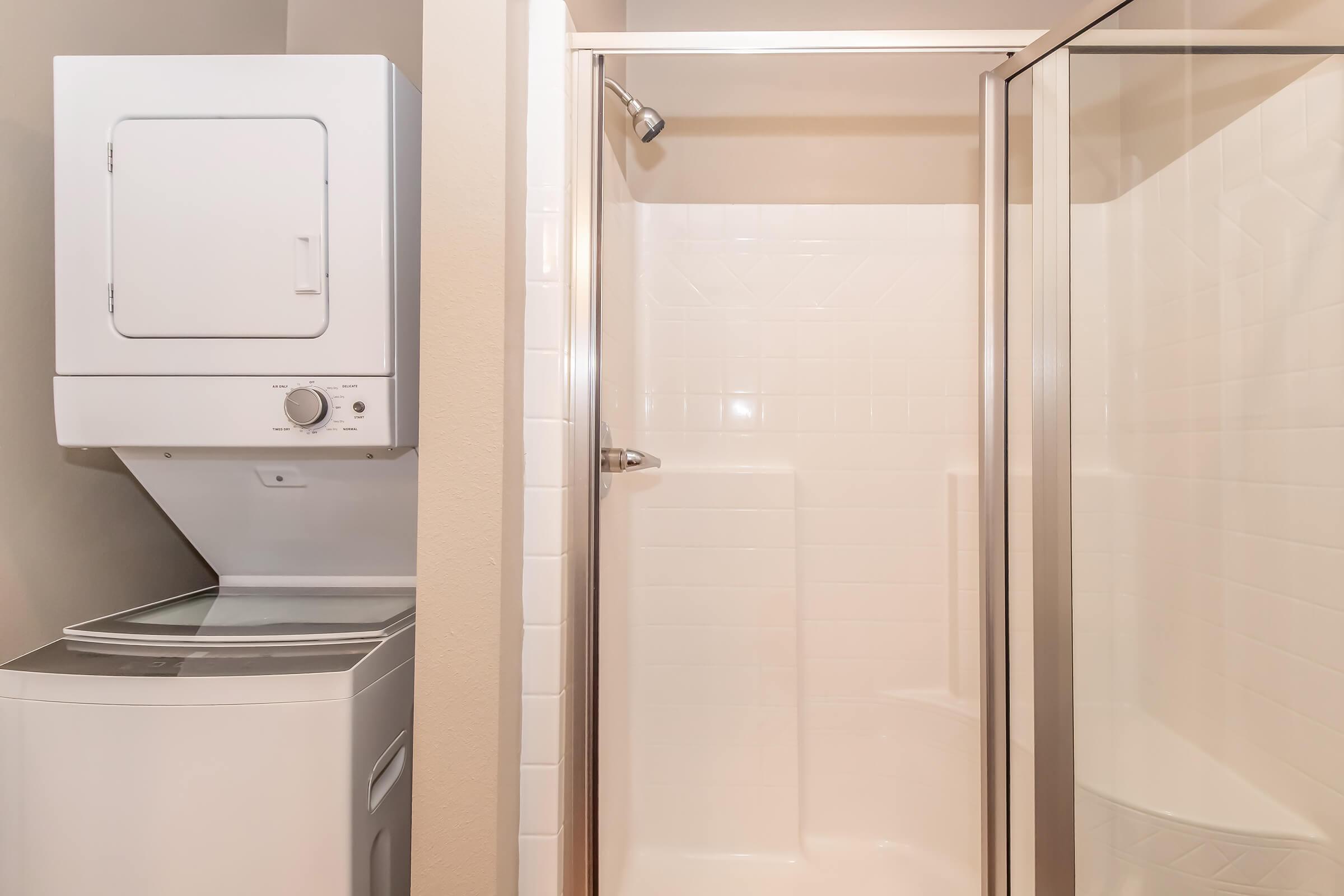 A compact laundry area featuring a stacked washer and dryer next to a glass-enclosed shower. The shower is tiled with white walls and has a silver showerhead, while the washer and dryer are located in a small nook, creating an efficient use of space.