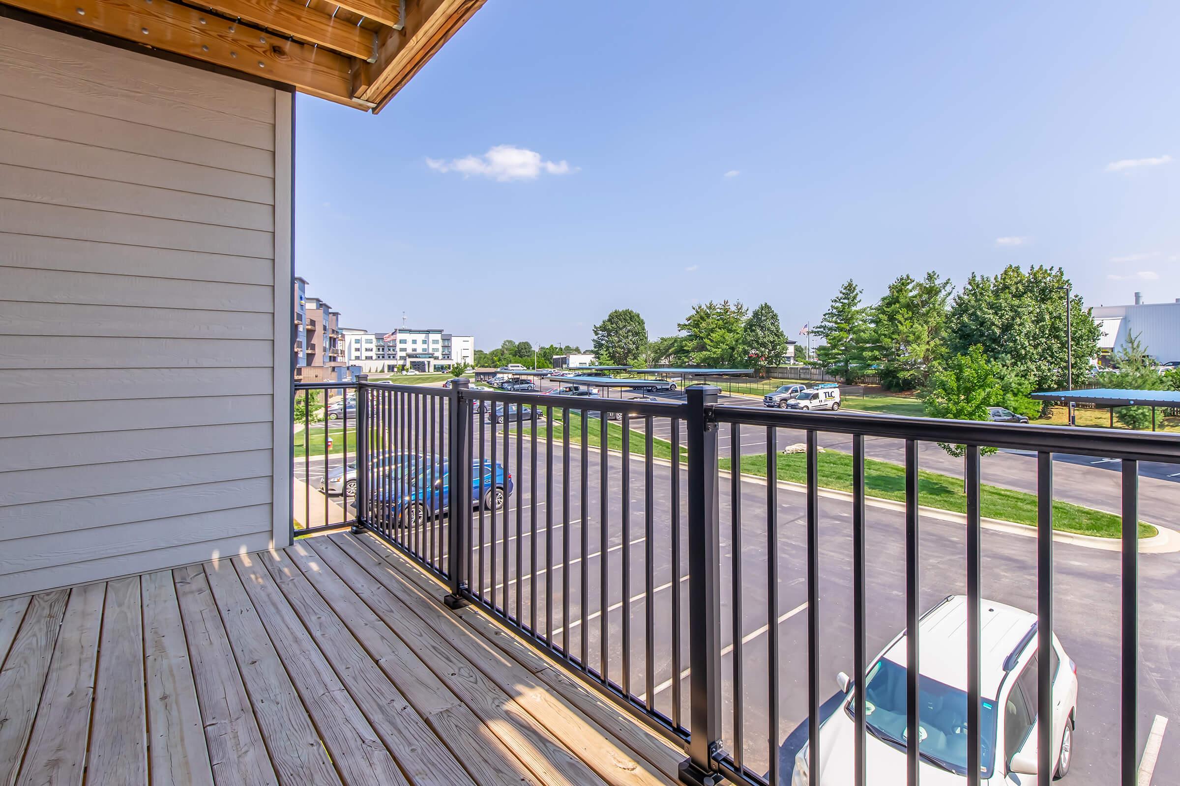 View from a balcony showcasing a wooden deck with a metal railing. In the background, there are trees, several parked cars, and buildings under a clear blue sky. The scene conveys a peaceful outdoor space with a suburban atmosphere.