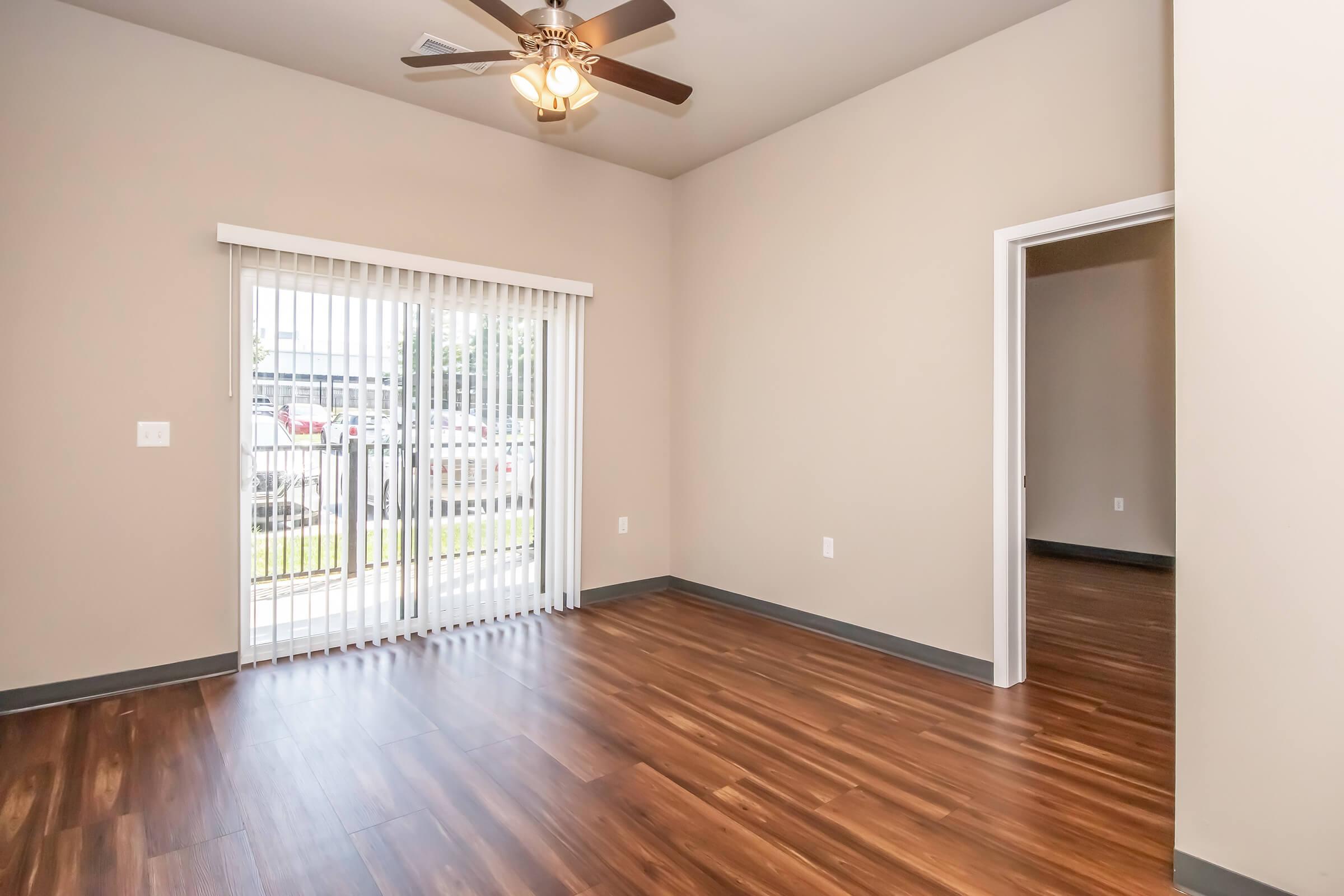 A spacious room with light brown walls and polished wood flooring. A ceiling fan is visible, and large sliding glass doors with vertical blinds lead to an outdoor area. A doorway on the right leads to another room, creating a bright and open layout.