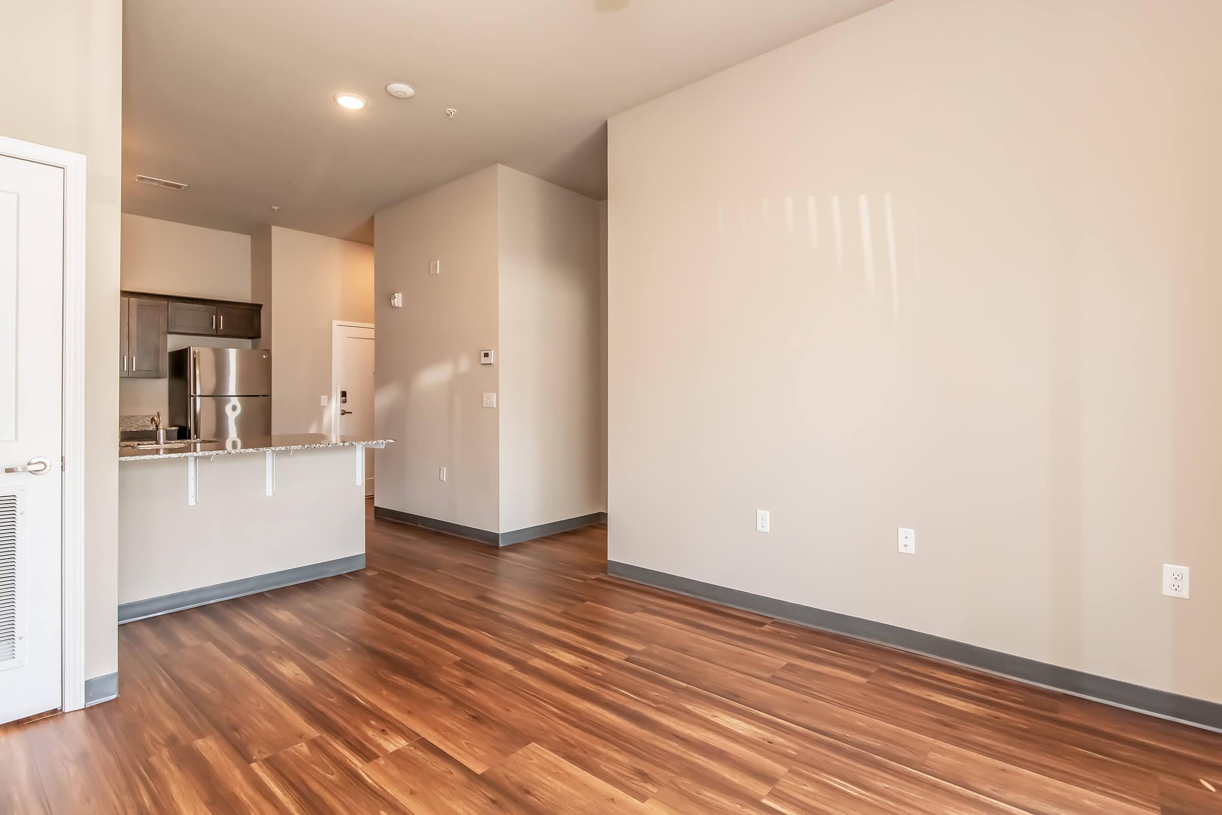 A spacious, newly designed apartment interior featuring light-colored walls and hardwood flooring. The room includes an open layout with a kitchen area visible in the background, equipped with modern appliances. Natural light may be coming from a window, creating a bright atmosphere.