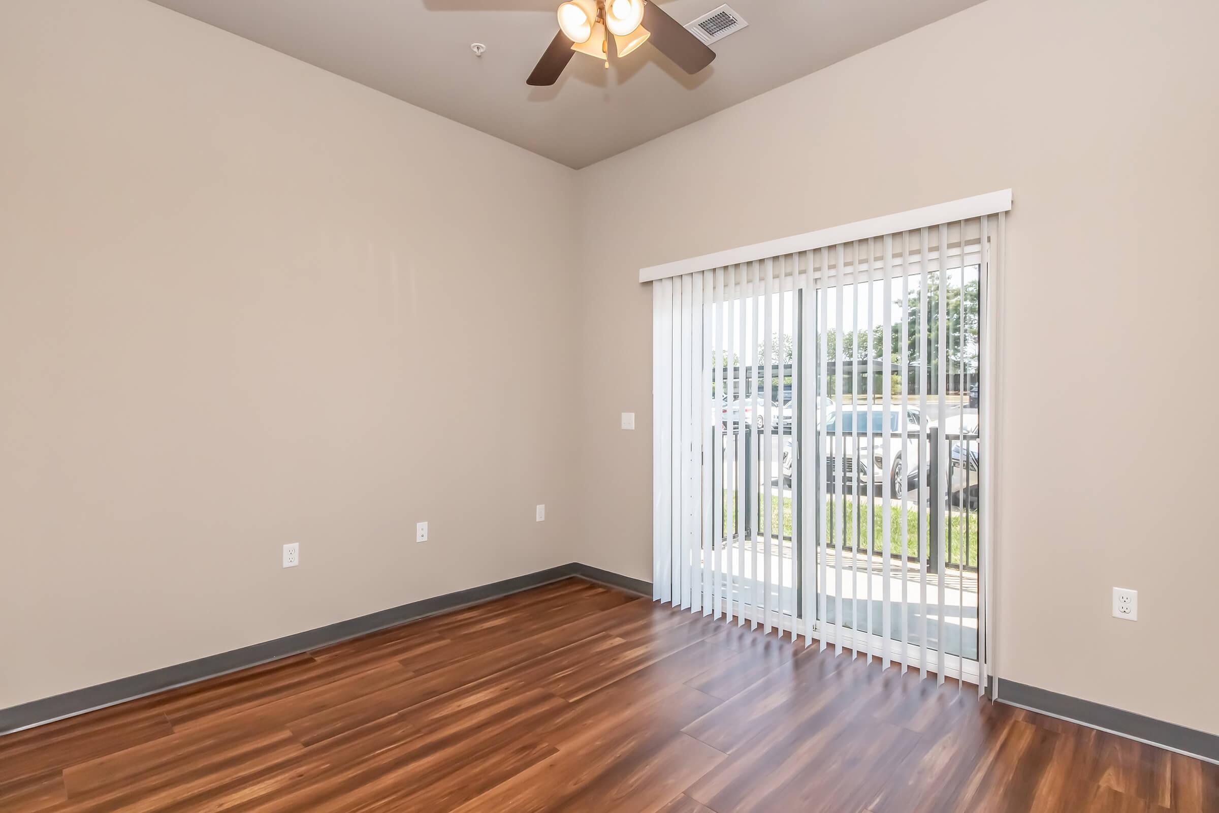 Empty room with wooden laminate flooring, beige walls, and a ceiling fan. There are vertical blinds covering a sliding glass door that leads to an outdoor area. The room has no furniture and features natural light from outside.
