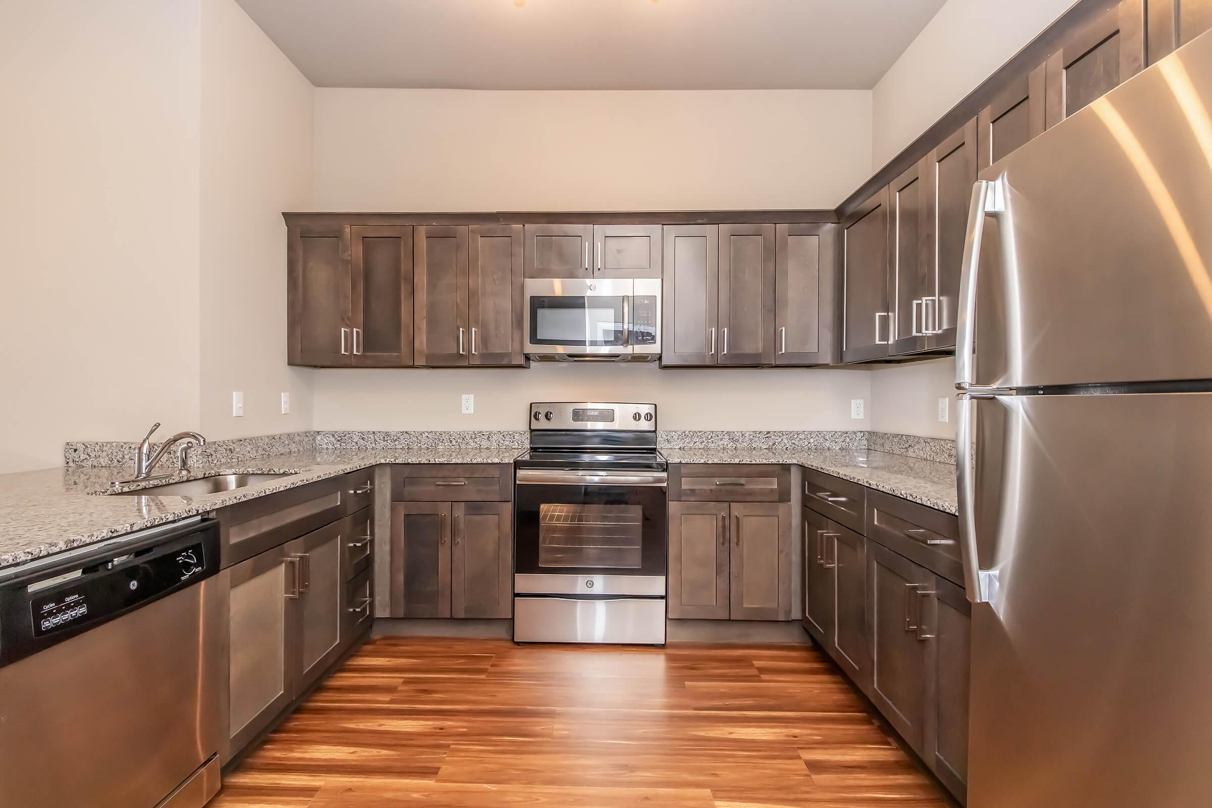 A modern kitchen featuring dark wooden cabinets, stainless steel appliances including an oven, microwave, and refrigerator, and a granite countertop. The floor is hardwood, and the space is well-lit with natural light.