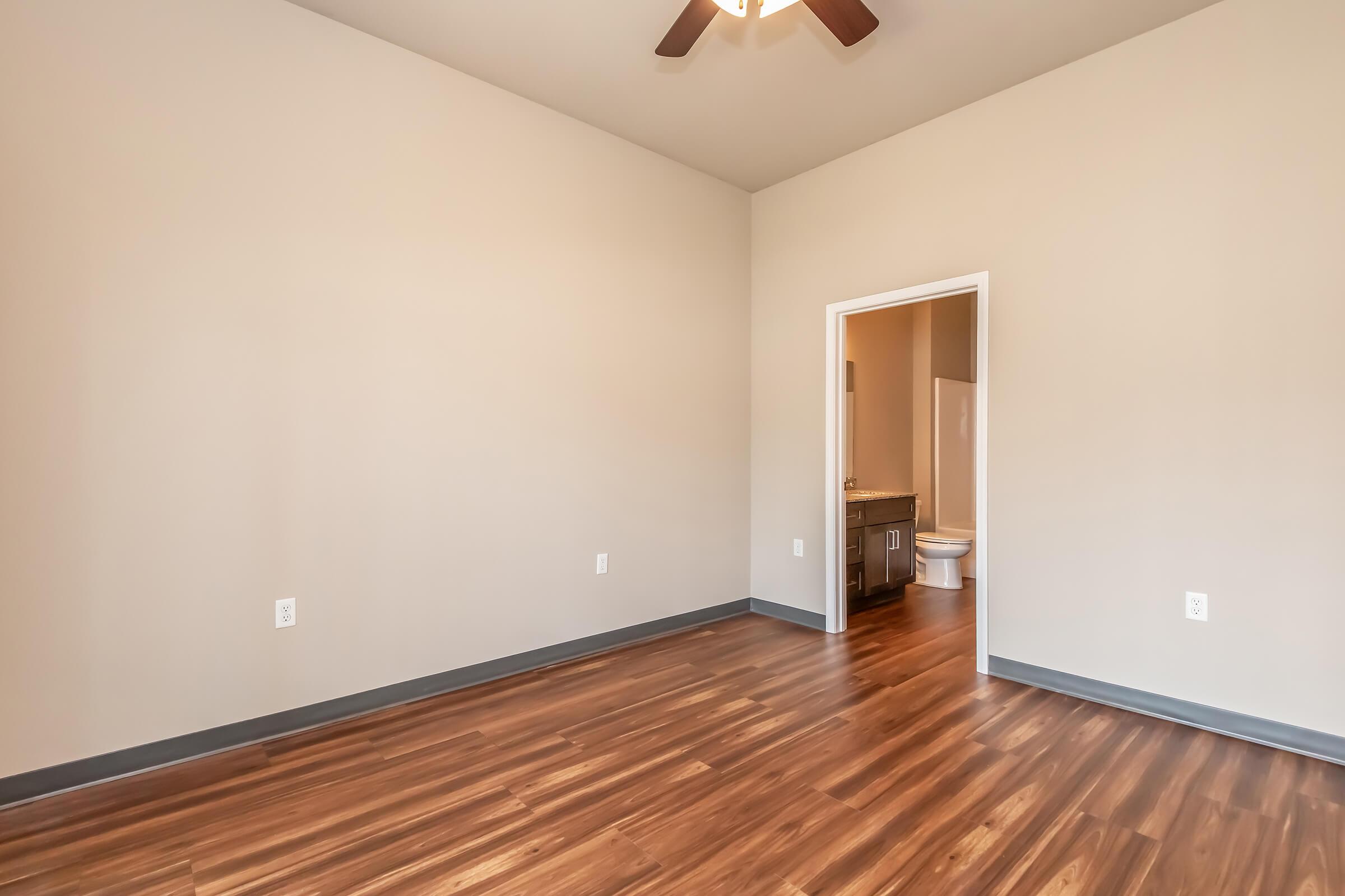 Empty room with light brown walls and a wooden floor. A ceiling fan is visible. A doorway leads to a bathroom, which is not fully visible. The space is bright and minimalistic, with no furniture present.