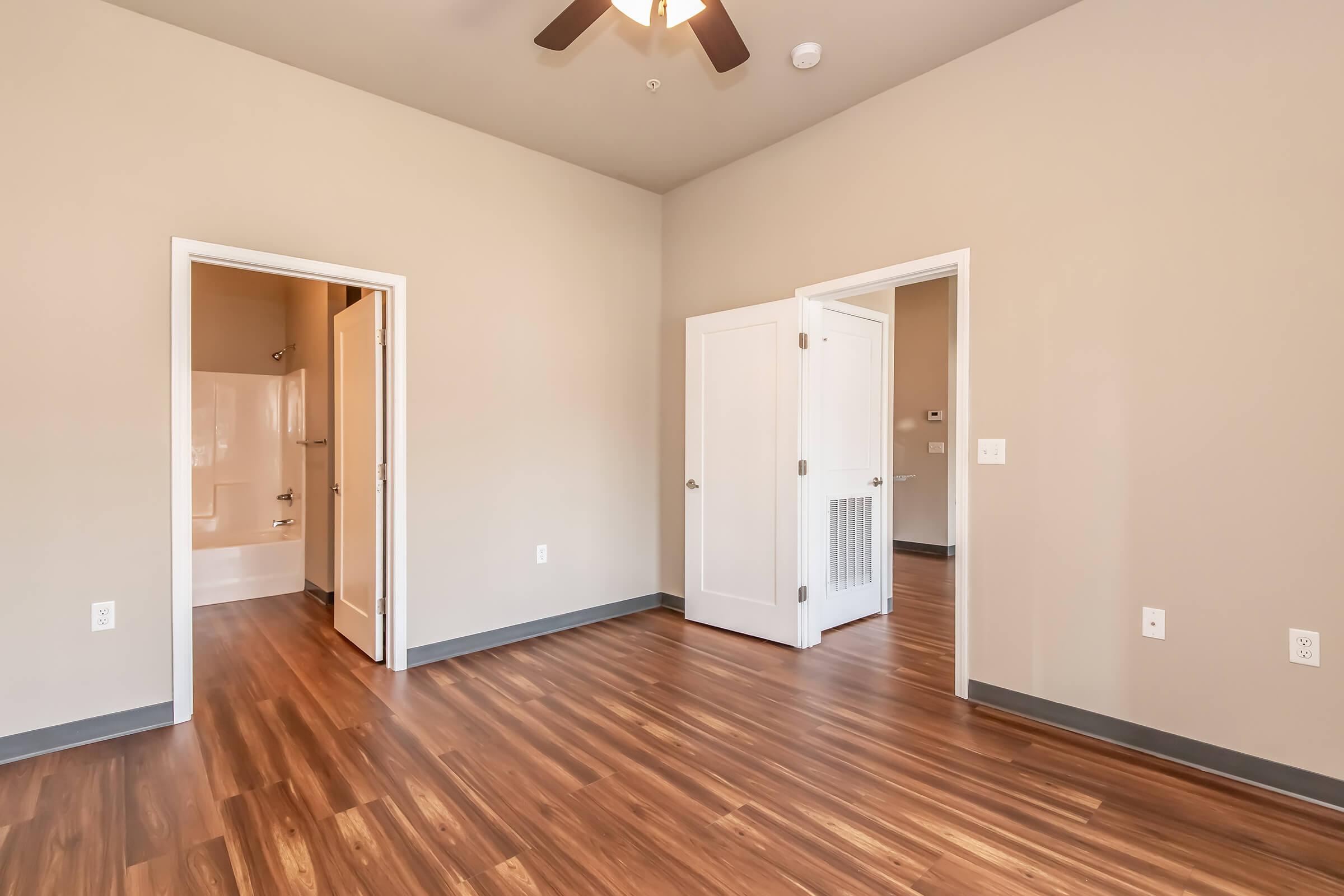A spacious, empty room with light brown walls and hardwood floors. There are two doorways leading to adjacent spaces, and a ceiling fan is visible overhead. The room is well-lit, with natural light coming from unseen windows. A bathroom area is partially visible through one of the doors.