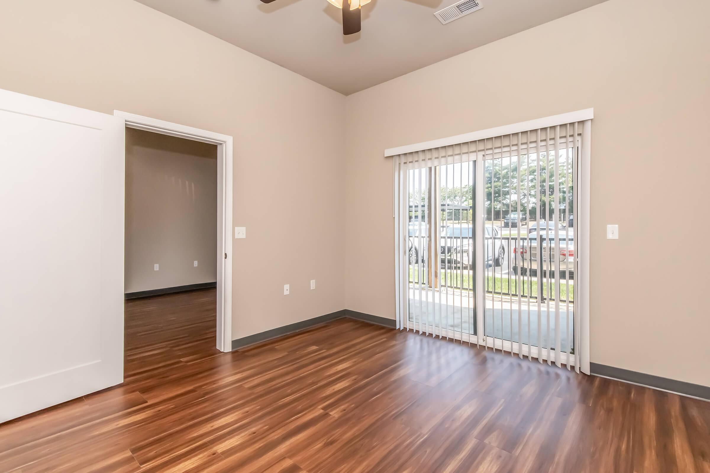 A spacious room featuring hardwood-style flooring, a ceiling fan, and a sliding glass door with vertical blinds that leads to an outdoor area. There is an adjoining room visible in the background. The walls are painted a neutral color, creating a bright and airy atmosphere.