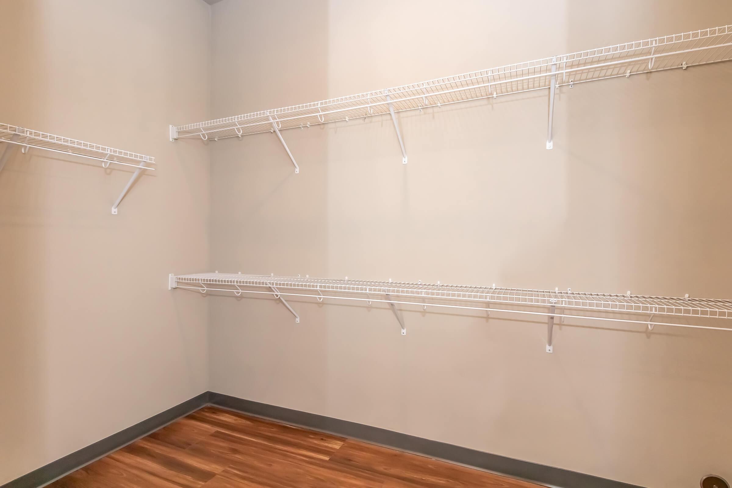 Empty closet with no doors, featuring white wire shelving on the walls and a wooden floor. The walls are painted a neutral color, and the shelves are positioned at various heights. The space is well-lit and organized, suitable for storage or personal items.