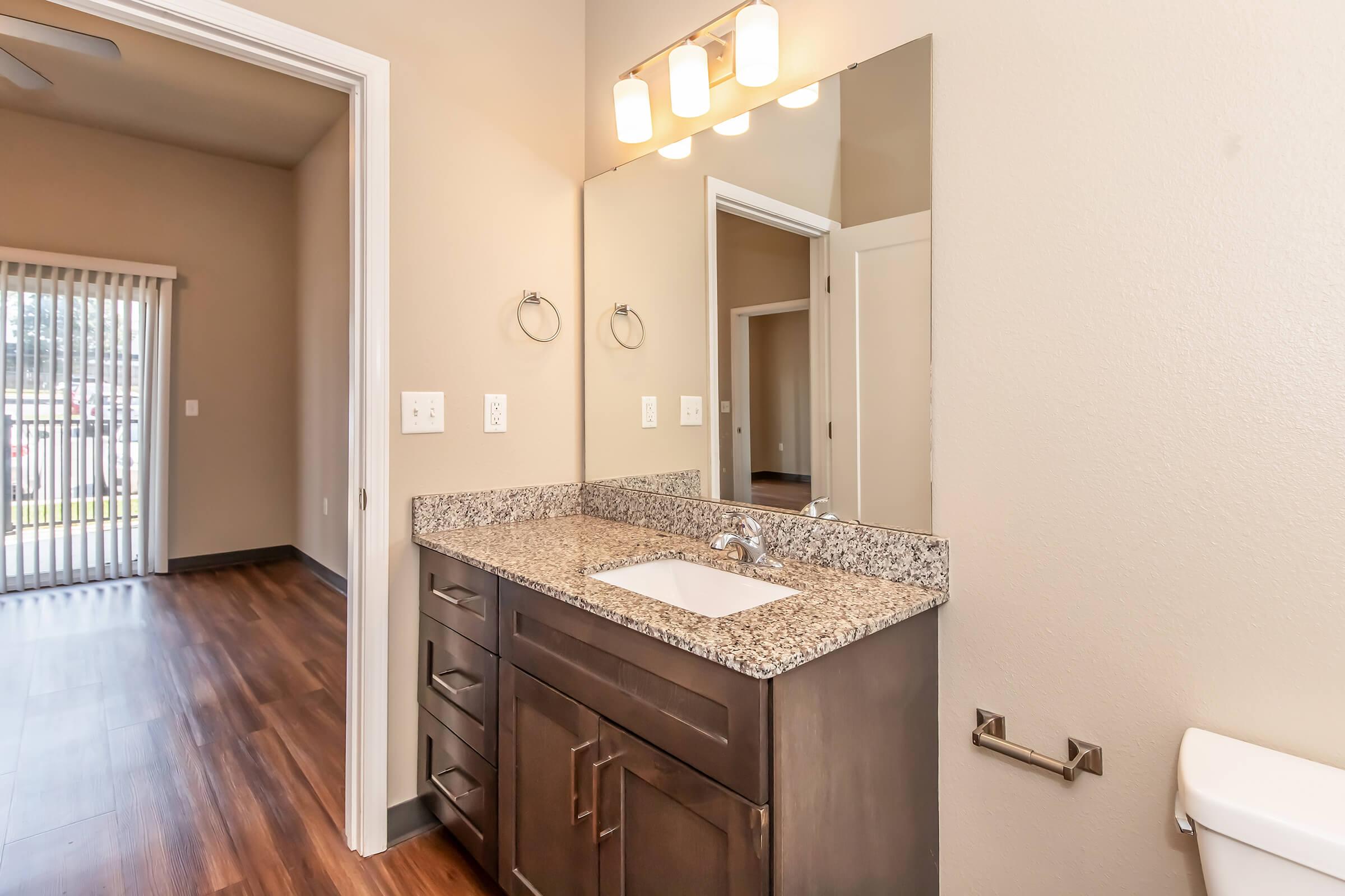 A bathroom with a modern double sink vanity featuring a granite countertop, a large mirror above, and stylish light fixtures. The room has light brown walls and dark wood flooring, with a doorway leading to a spacious area visible in the background. A white toilet is positioned against one wall.