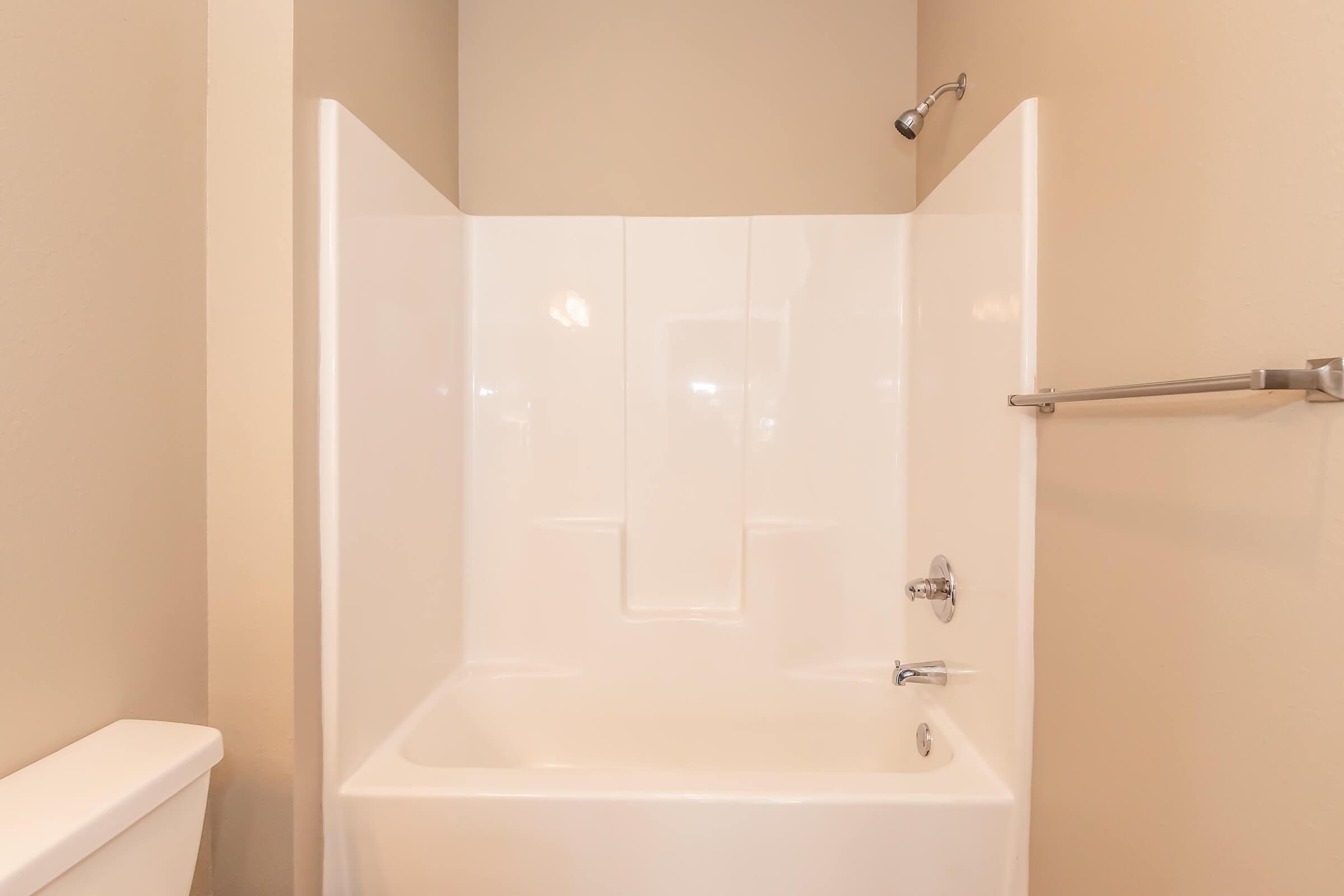 A clean, modern bathroom featuring a white shower and bathtub combination. The walls are painted in a neutral tone, and there is a showerhead mounted above. A towel bar is visible on the side, with a toilet partially seen in the foreground. The overall design is simple and functional.