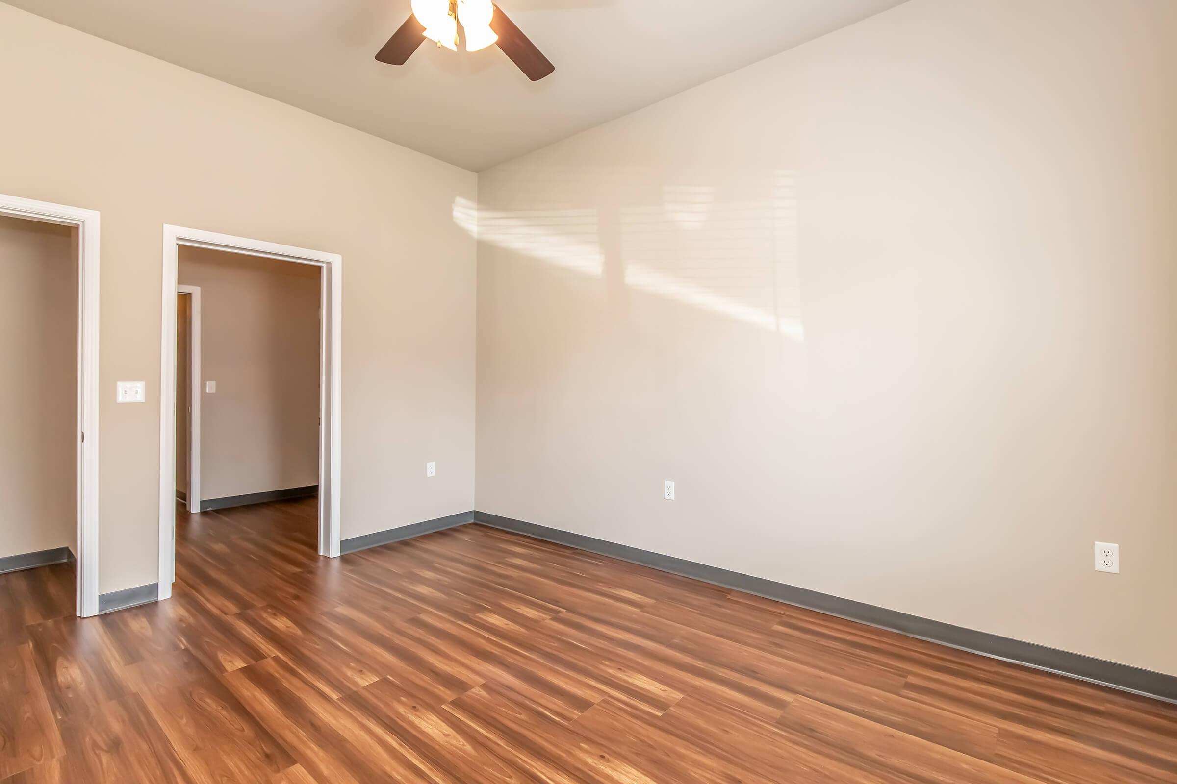 Empty room with a wooden floor and light-colored walls. A ceiling fan is visible, and there are two doorways leading to other areas. Natural light is streaming in from a window, casting soft shadows on the walls. The overall ambiance is bright and spacious, ideal for an apartment or living space.