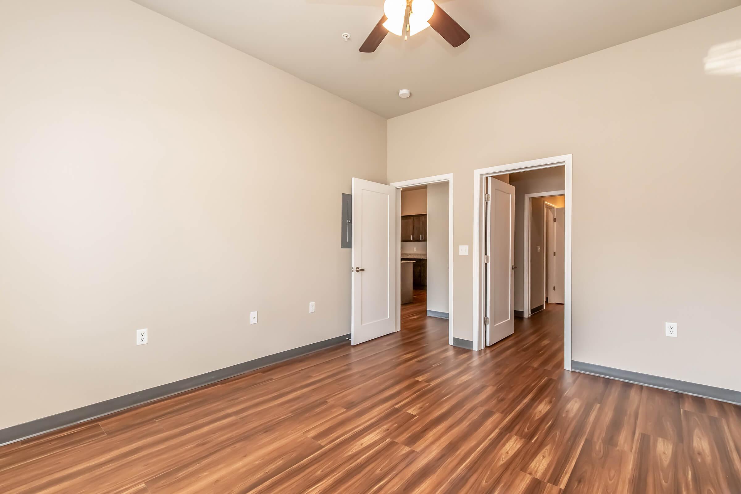 A spacious room featuring light beige walls, a ceiling fan, and dark wood-like flooring. Two open doors lead to adjacent areas, while a third door is partially visible on the left. The overall aesthetic is modern and inviting, creating a bright and airy atmosphere.