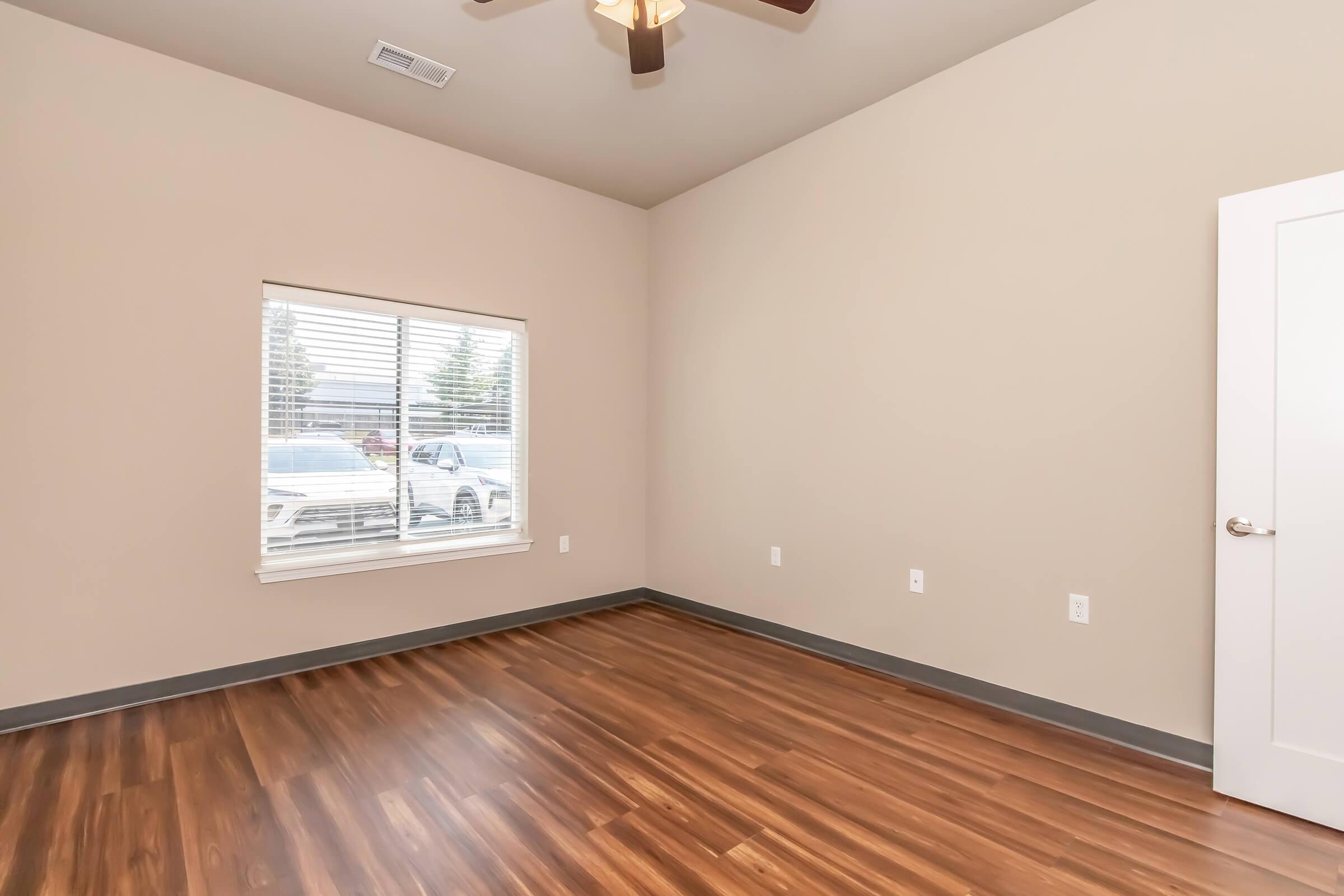 A vacant room with light-colored walls and a wooden floor. A window with blinds lets in natural light, showing a view of a vehicle outside. There is a ceiling fan and a single door leading out of the room. The space is clean and modern, suggesting potential for various uses.