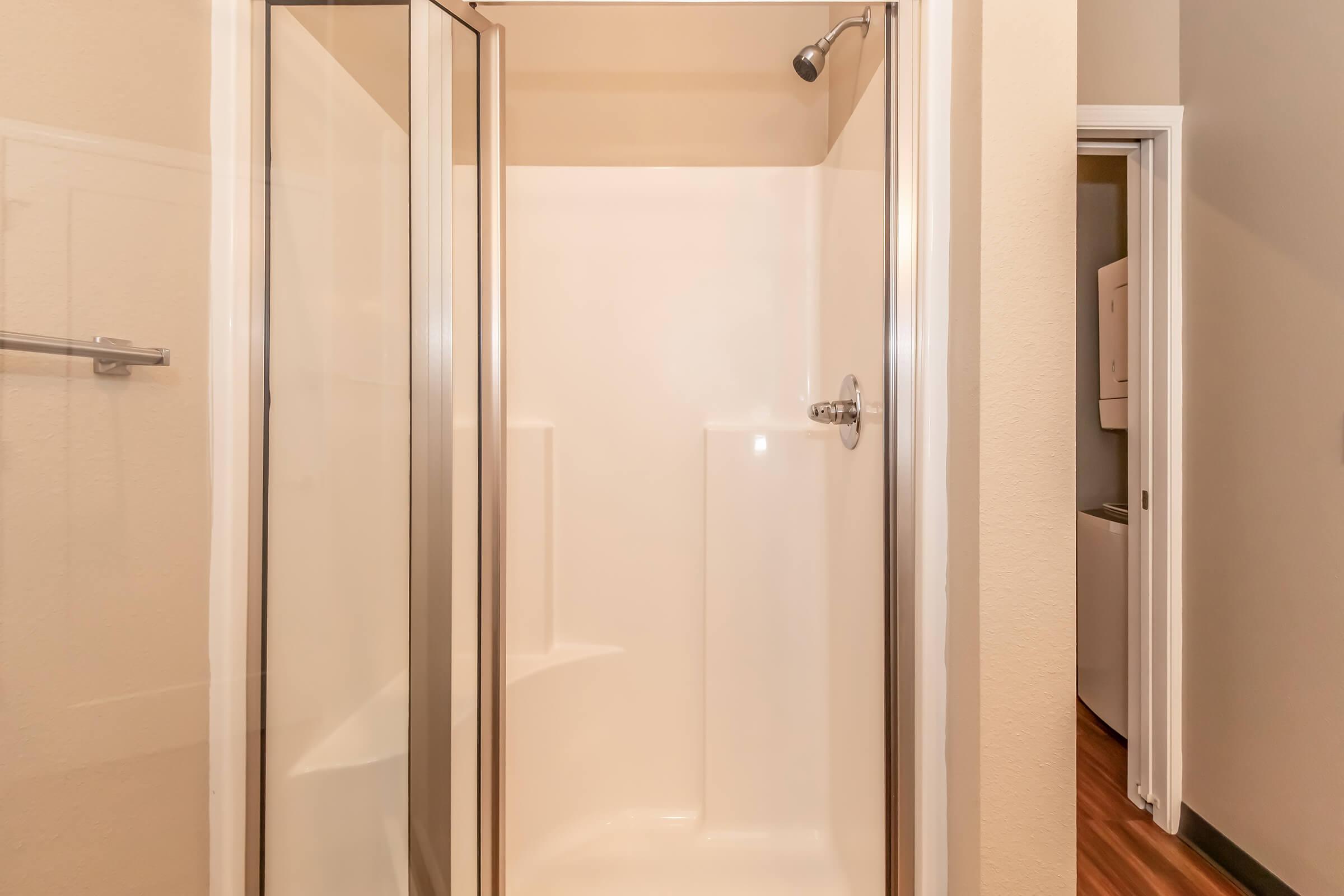 A clean and empty bathroom shower with glass doors, featuring a chrome showerhead and built-in shelving on the walls, illuminated by soft overhead lighting. The surroundings have light-colored walls, creating a fresh and modern atmosphere.