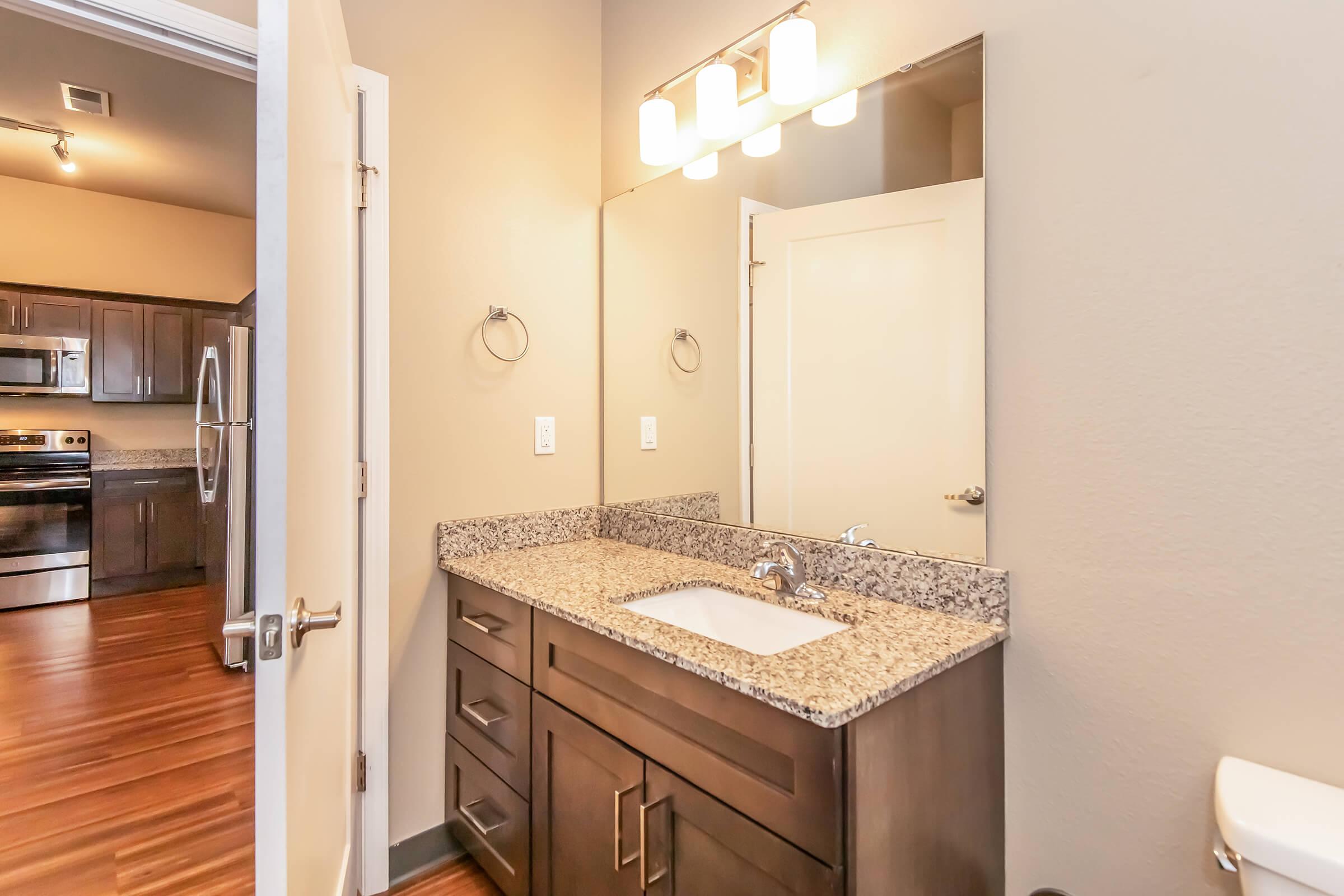 A modern bathroom featuring a granite countertop with a sink, a large mirror above, and sleek cabinetry. The walls are painted in a neutral tone, and there is a towel ring on the wall. A doorway leads to another room, while the flooring is a warm wood finish. Modern lighting fixtures are mounted above the mirror.