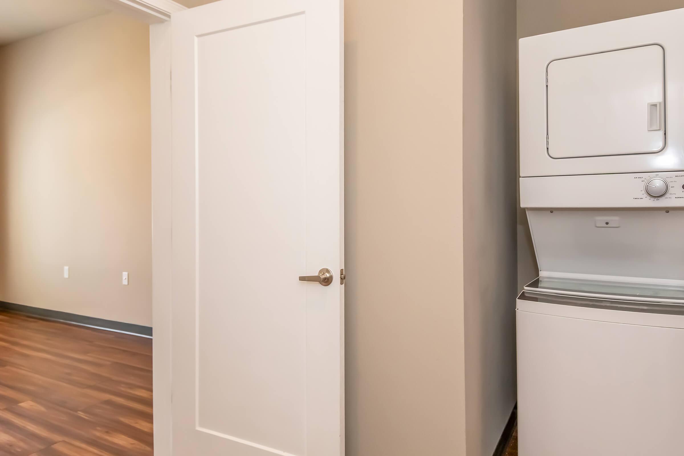 A laundry area featuring a stacked washer and dryer against a beige wall, with a white door partially visible on the left. The room has laminated wood flooring and a spacious feel, suitable for a residential setting.