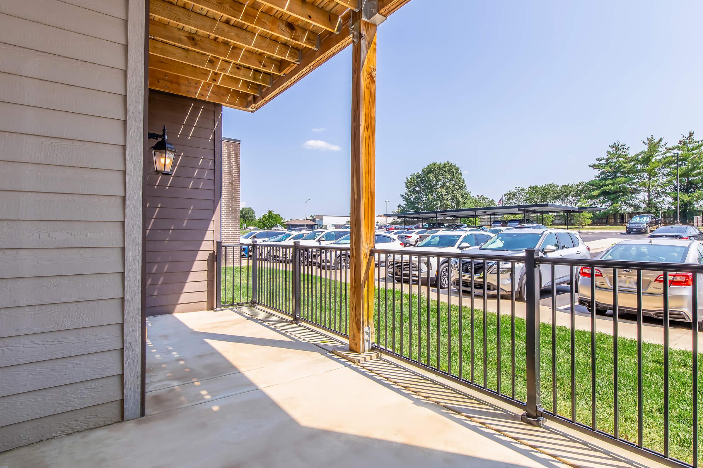A spacious balcony with wooden beams and a metal railing, overlooking a parking lot filled with cars. The clear blue sky and green grass enhance the area's inviting atmosphere. Sunlight casts soft shadows on the concrete floor, creating a warm and welcoming vibe.
