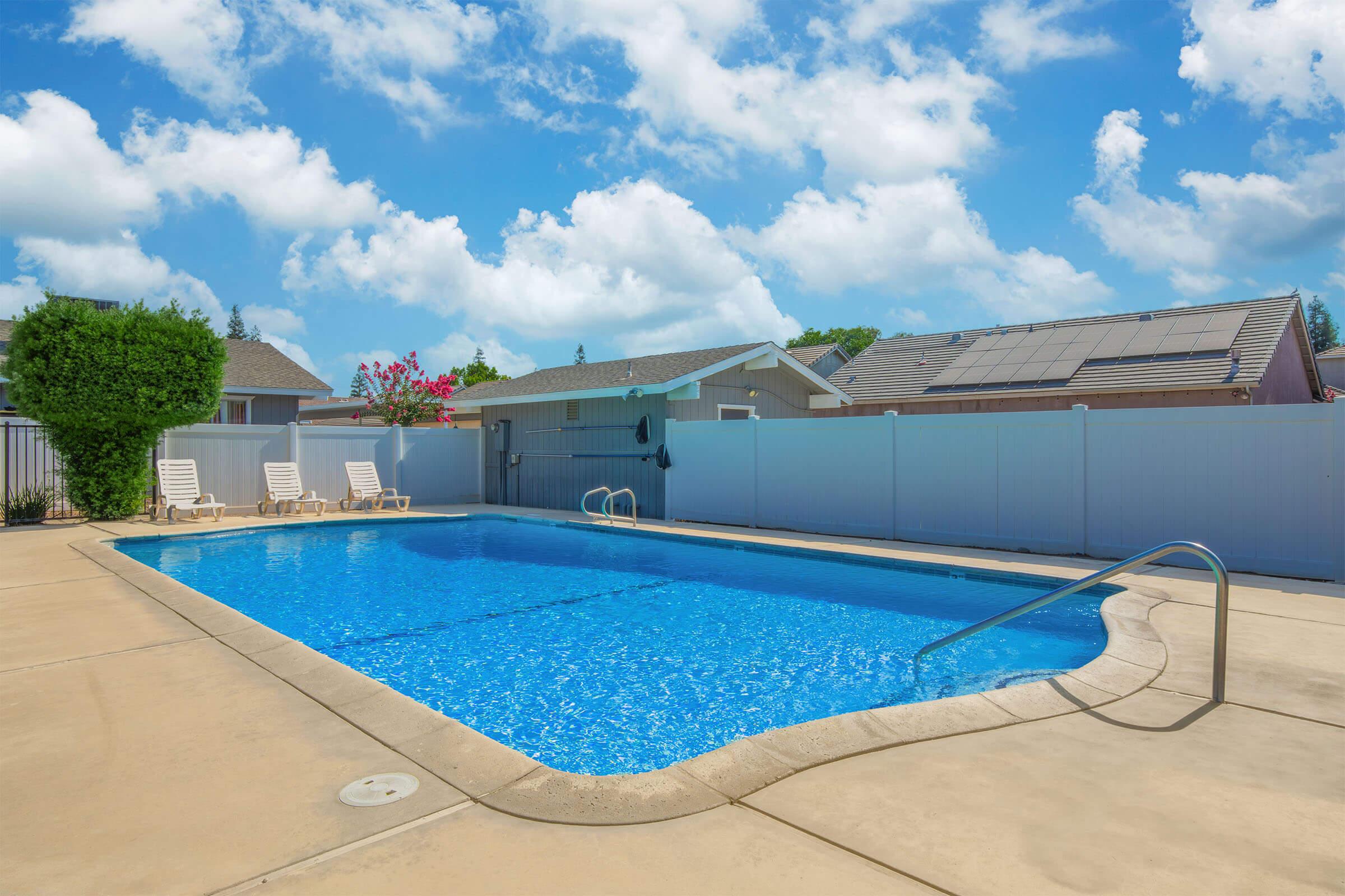 A clear blue swimming pool surrounded by a concrete patio, with several lounge chairs on one side. In the background, there are houses with solar panels and greenery. The sky is bright with scattered clouds, creating a sunny, inviting atmosphere.