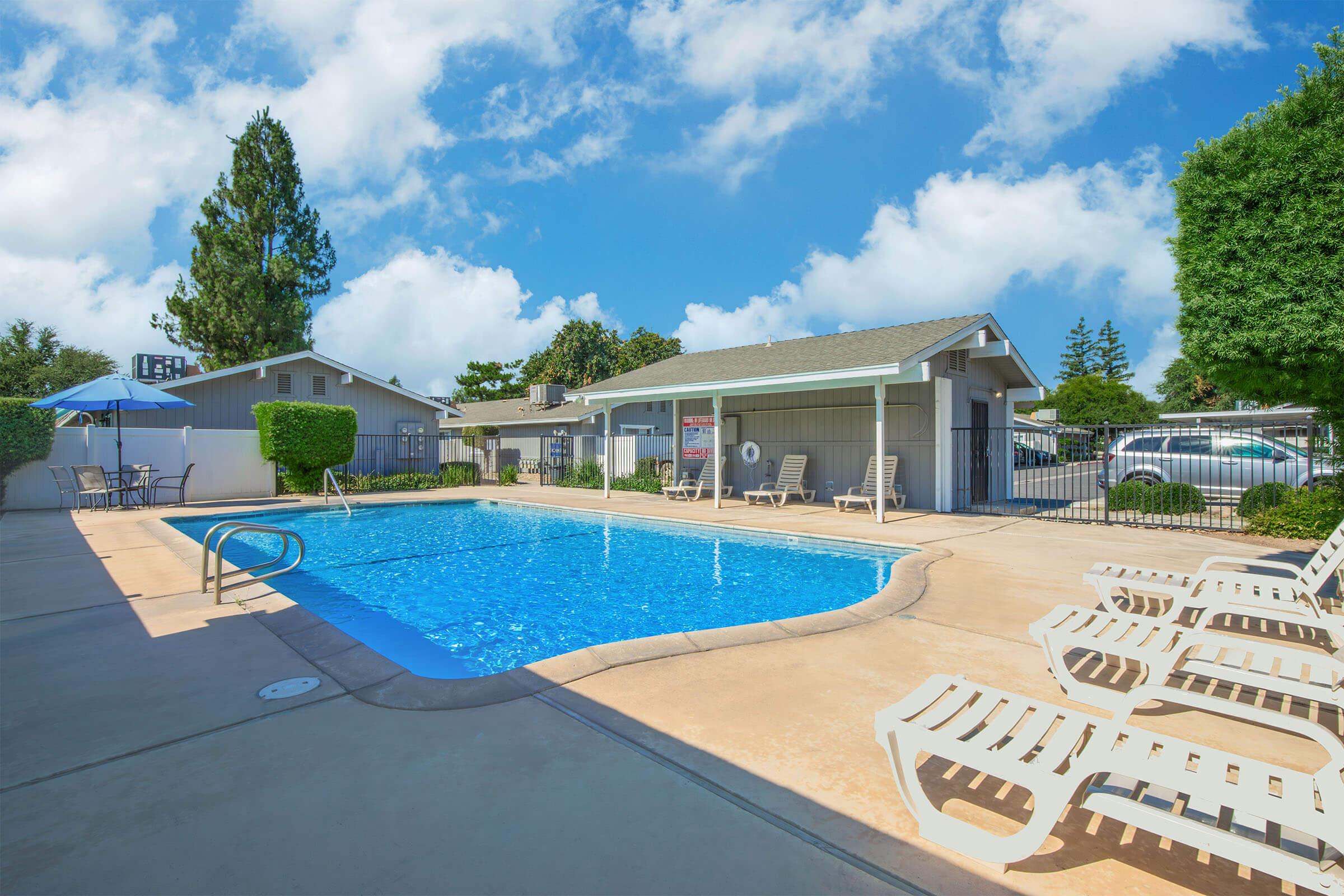 A bright and inviting outdoor swimming pool surrounded by lounge chairs and shaded areas. The pool area is bordered by well-maintained landscaping, including trees and shrubs, and a light blue sky with fluffy clouds overhead. There is a nearby building with a covered area for relaxation.