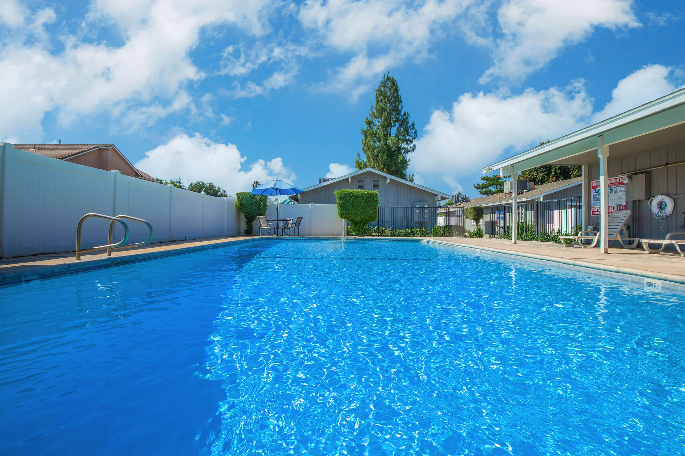 A clear blue swimming pool with a ladder, surrounded by green hedges and lounge chairs. In the background, there are several buildings and a bright blue sky with fluffy white clouds. An umbrella provides shade near the pool area, creating a relaxing outdoor atmosphere.