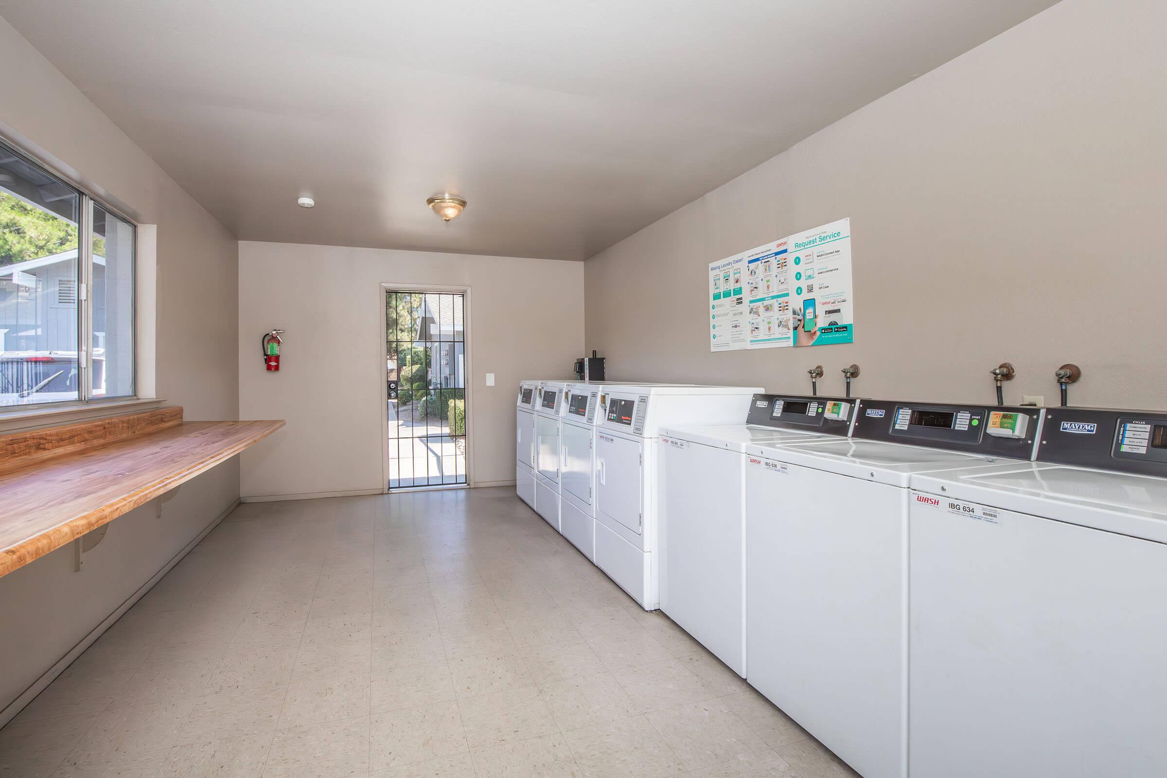 Interior view of a laundry room featuring several white washing machines lined up against a wall. A countertop made of wood runs along one side, and there is a door leading outside. A poster with laundry instructions is visible on the wall, along with a fire extinguisher near the entrance.