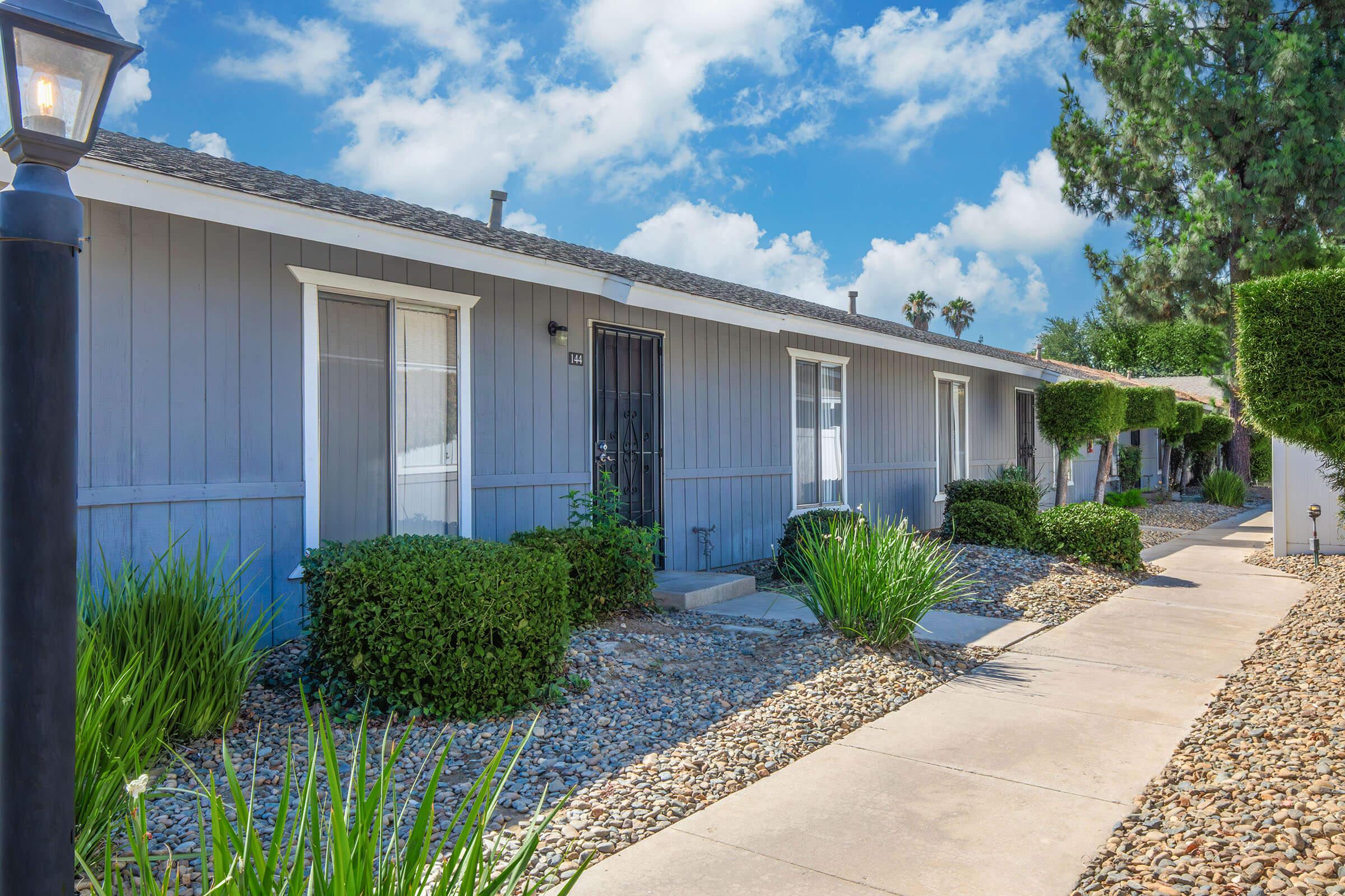 A row of single-story gray buildings with white windows and doors, lined with neatly trimmed shrubs and rocks. A walkway leads to the entrance of each unit, framed by tall trees and a bright blue sky with scattered clouds.