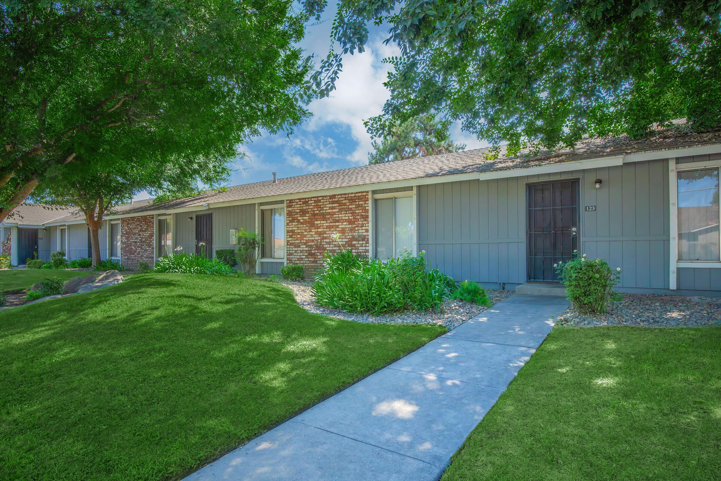 A row of residential buildings with a manicured lawn, green shrubs, and trees. The pathway leads to the front doors of the homes, each featuring a small porch. The sky is clear with a few clouds, creating a bright and inviting atmosphere.