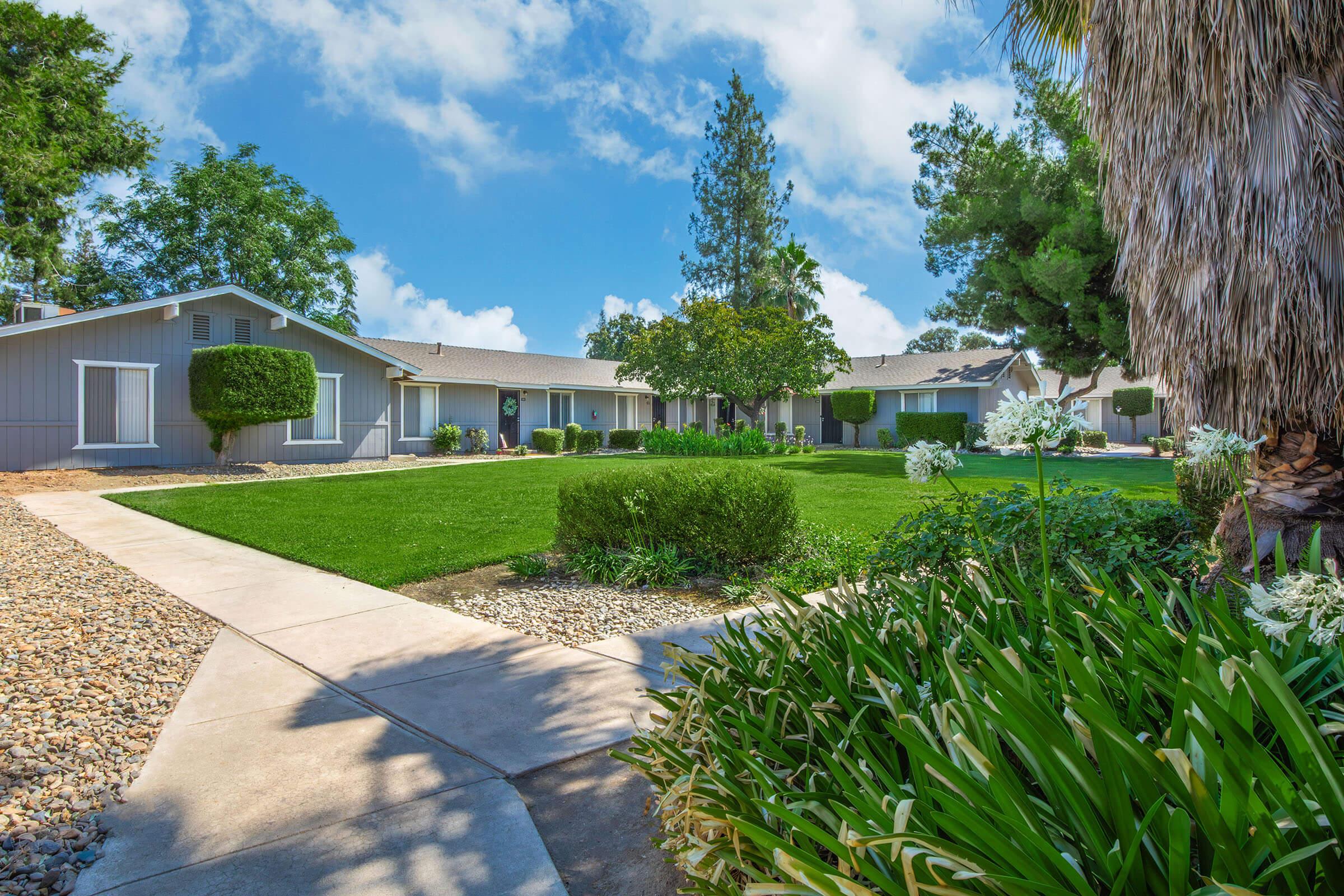 A well-maintained residential landscape featuring a pathway leading to several single-story gray houses. Lush green grass surrounds the homes, with neatly trimmed shrubs and trees providing shade. Bright blue skies and scattered clouds create a serene atmosphere.
