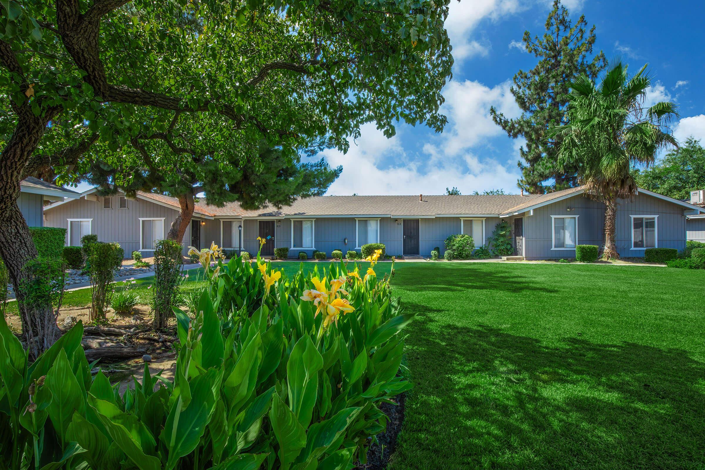 A well-maintained single-story building surrounded by lush green grass and colorful flowering plants. Trees provide shade, and the sky is partly cloudy, creating a serene atmosphere. The structure features multiple windows, indicating a welcoming residence or community space.