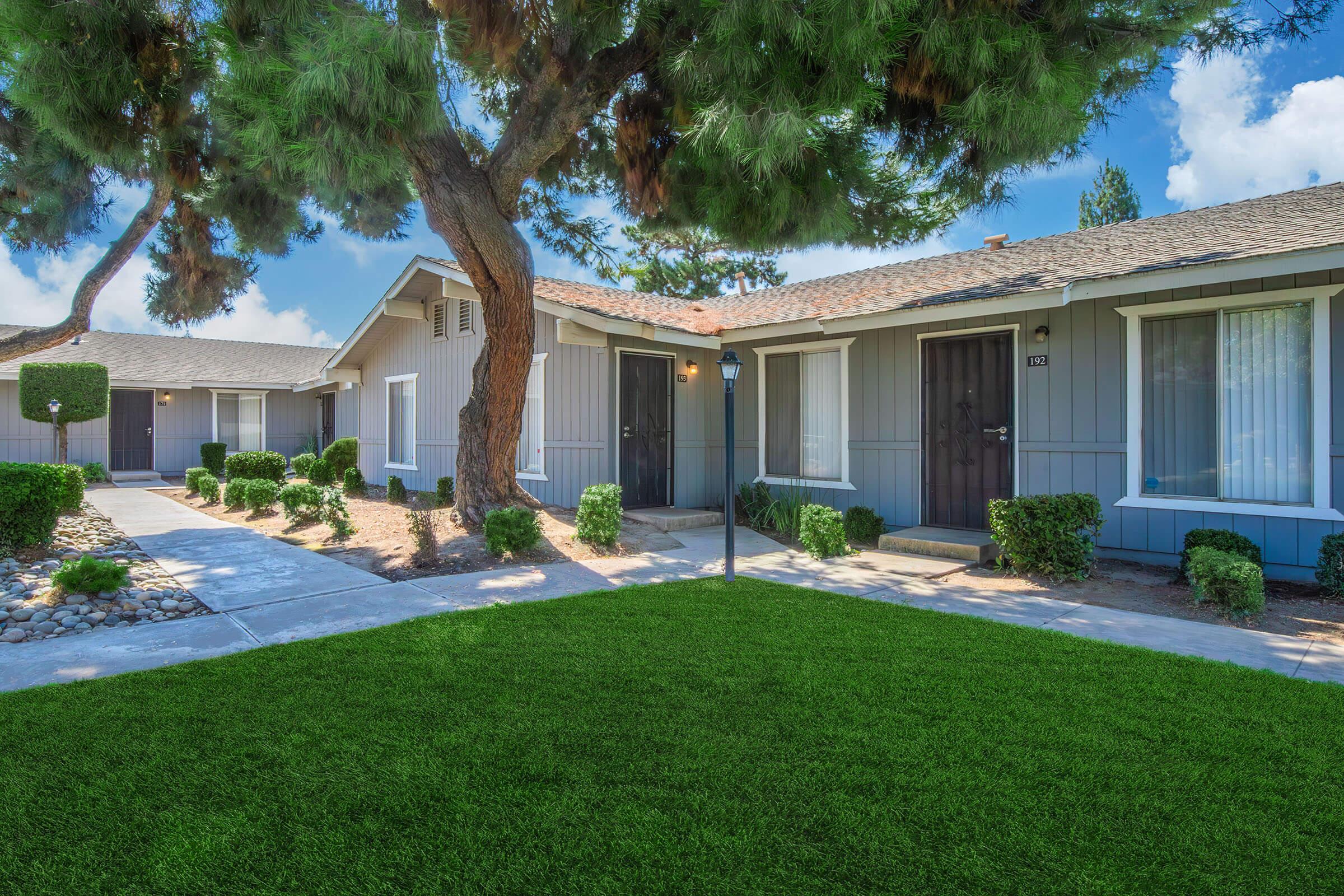 A well-maintained exterior of a residential area featuring gray houses with dark doors, neatly trimmed hedges, and a lush green lawn. A large tree provides shade, and a concrete pathway leads to the front doors. The sky is clear with a few clouds.