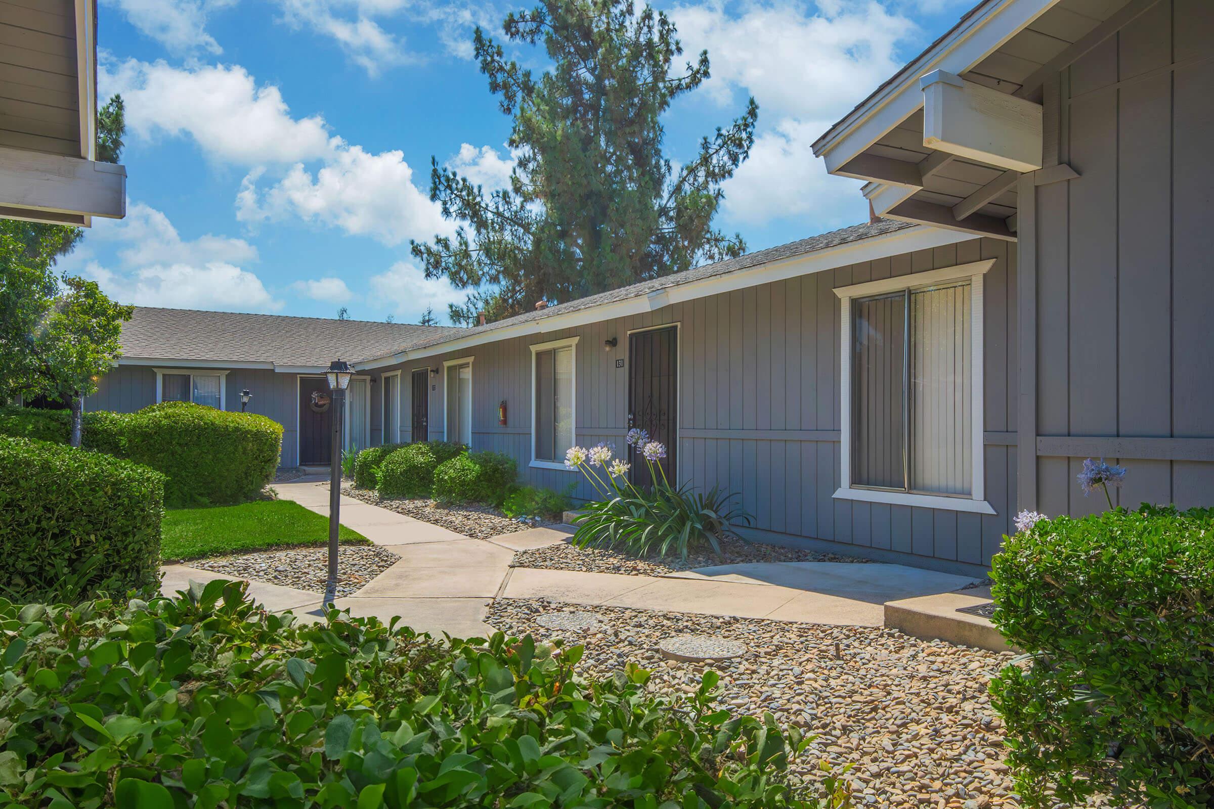 A landscaped pathway leads through a courtyard with neatly trimmed hedges and flowering plants, connecting several light gray buildings. The sky is bright with fluffy white clouds, creating a serene and inviting atmosphere.