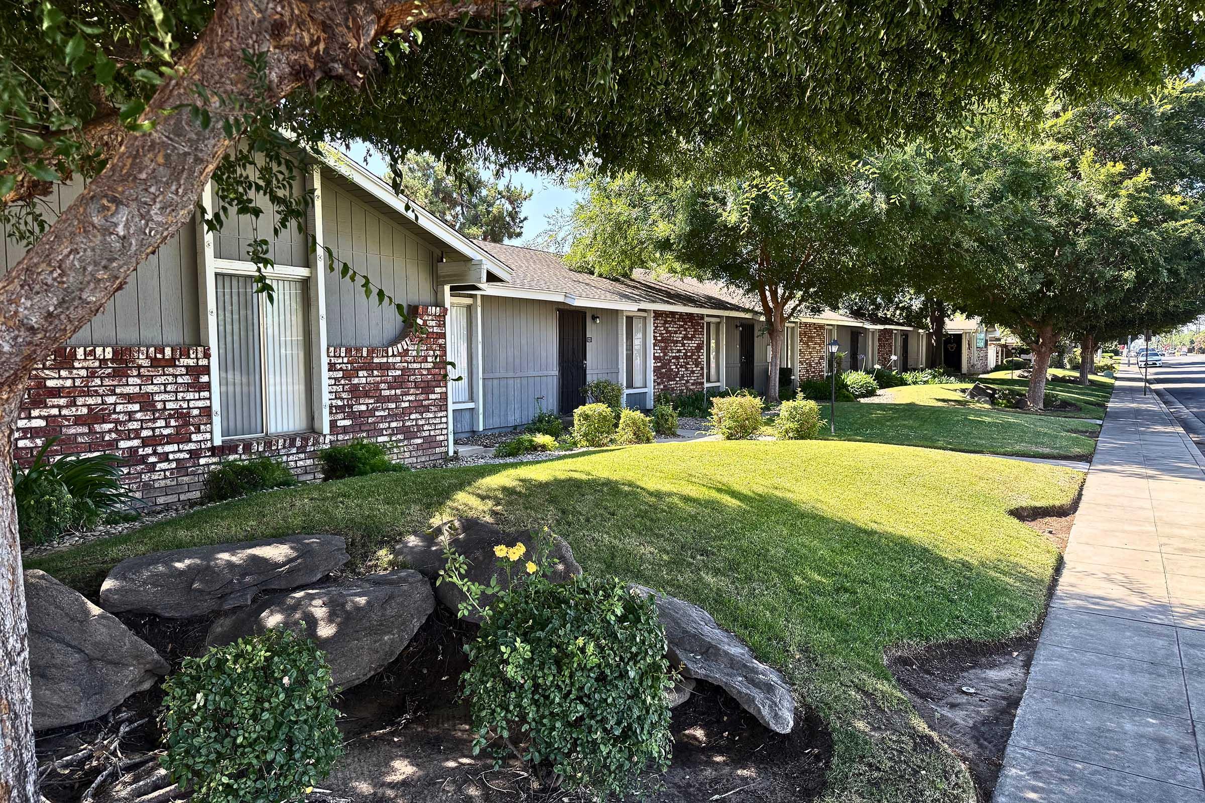 A row of single-story houses with brick facades and green lawns, bordered by shrubs and rocks. Tree-lined street with well-maintained landscaping, creating a peaceful suburban atmosphere.