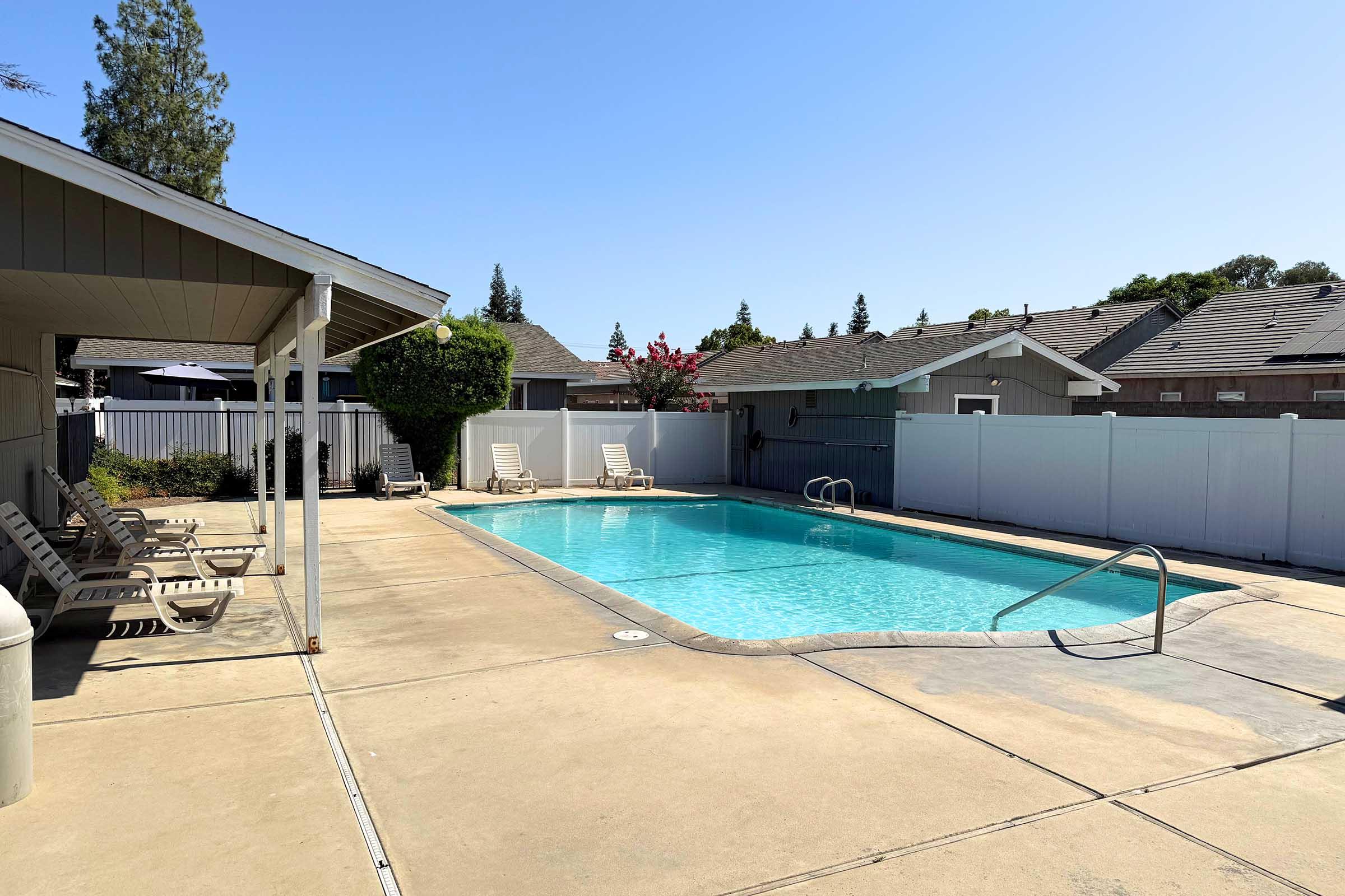 A clear blue swimming pool surrounded by a concrete patio and lounge chairs. A small shelter is visible on one side, with houses and trees in the background. The sky is clear and sunny, indicating a warm day. The pool area is enclosed by a white fence.