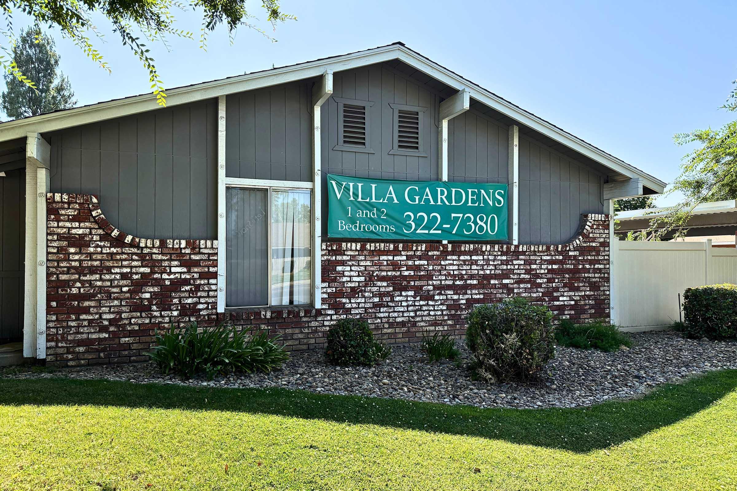 A residential building featuring a sign that reads "VILLA GARDENS" showcasing available 1 and 2-bedroom units, with a contact number "322-7380." The structure has a brick exterior and a well-maintained lawn with shrubs and decorative landscaping.