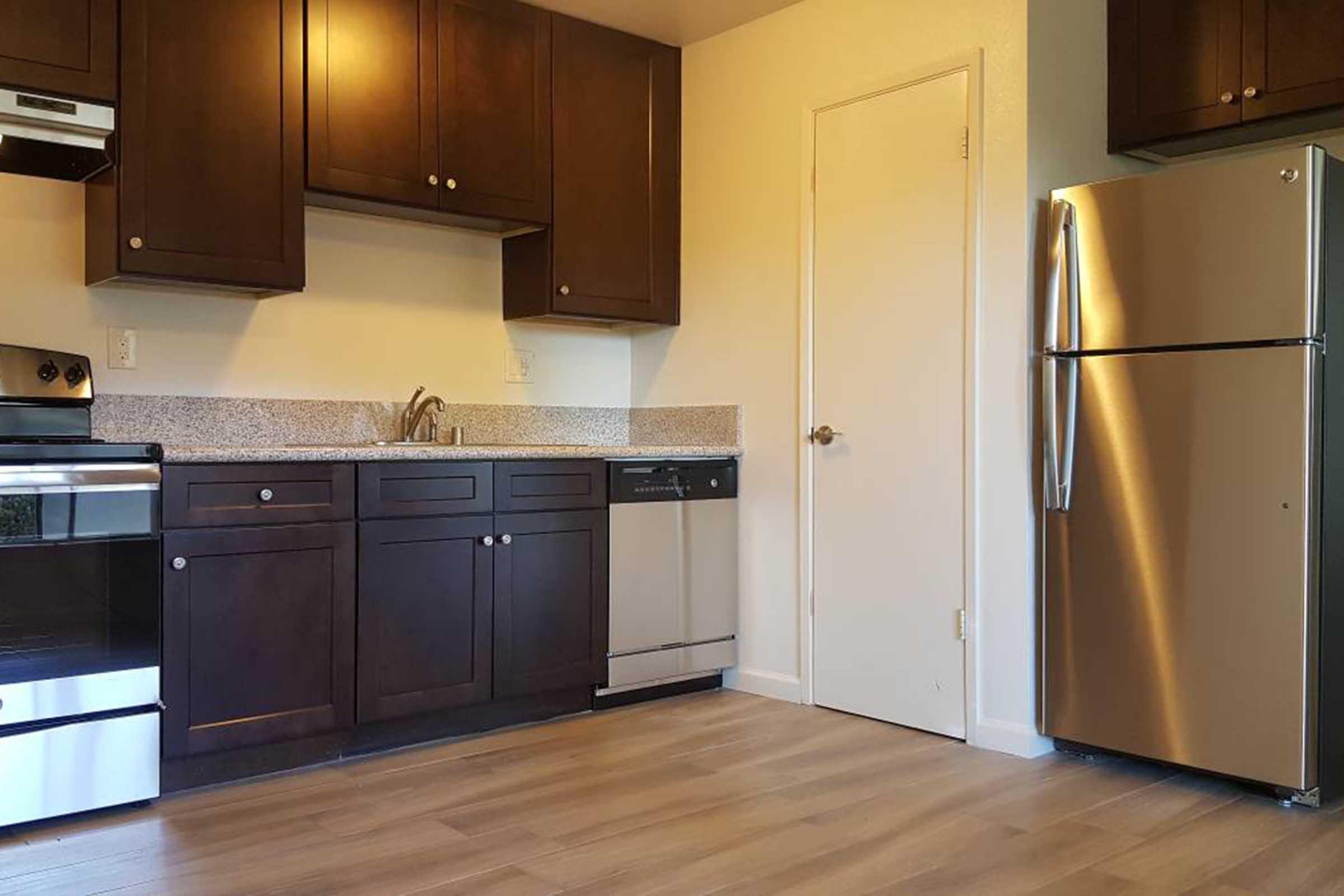 A modern kitchen featuring dark wooden cabinets, a countertop, a stainless steel refrigerator, a dishwasher, and an oven. The floor is made of light-colored wood, and there is a plain white door on one wall, creating a clean and functional space.