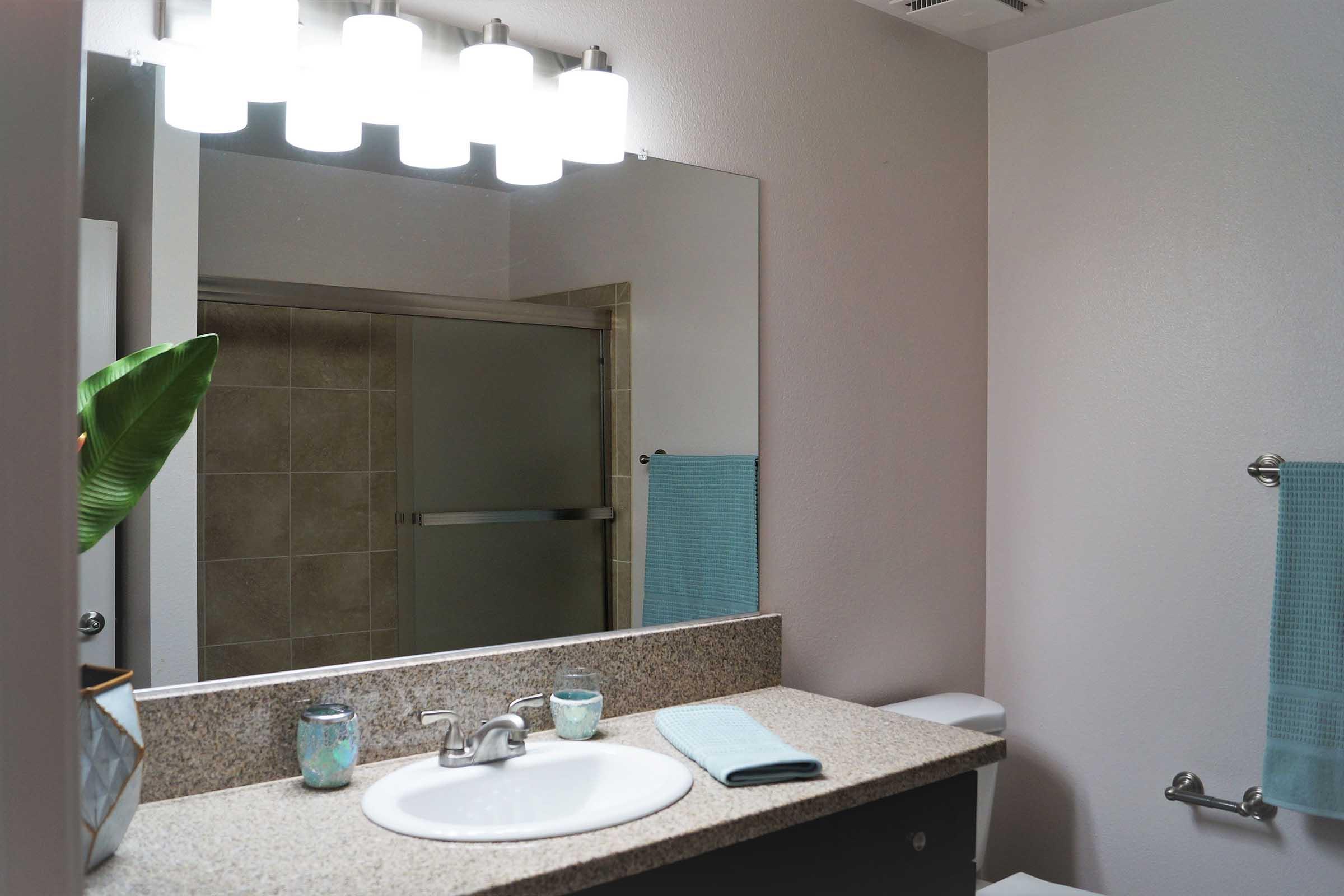 A modern bathroom featuring a granite countertop with a sink, two decorative candles, and a light fixture with four bulbs above the mirror. A shower enclosure is visible in the background alongside a towel rack with a light blue towel. A potted plant adds a touch of greenery to the decor.