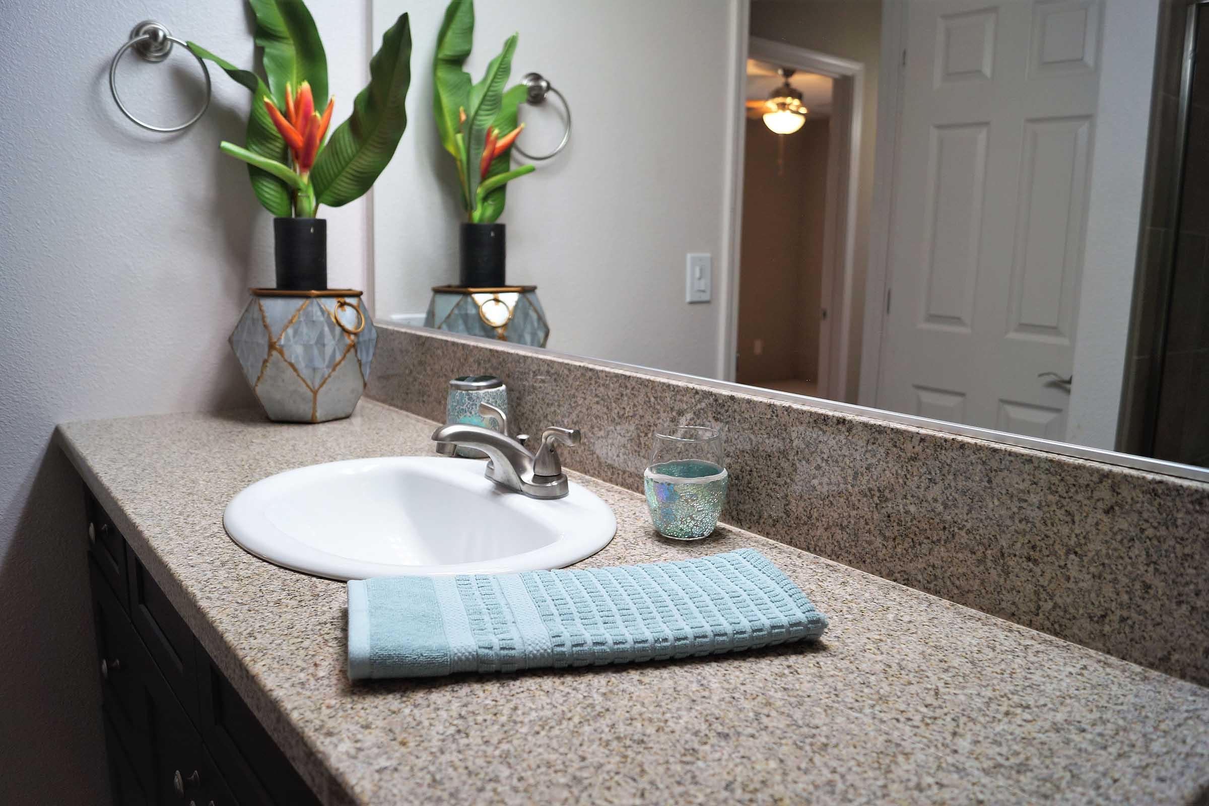 A well-lit bathroom with a granite countertop featuring a white sink. A light blue towel is neatly placed on the surface, along with a small decorative glass and a potted plant with vibrant green leaves and orange flowers. A mirror reflects the scene, enhancing the space's brightness and elegance.