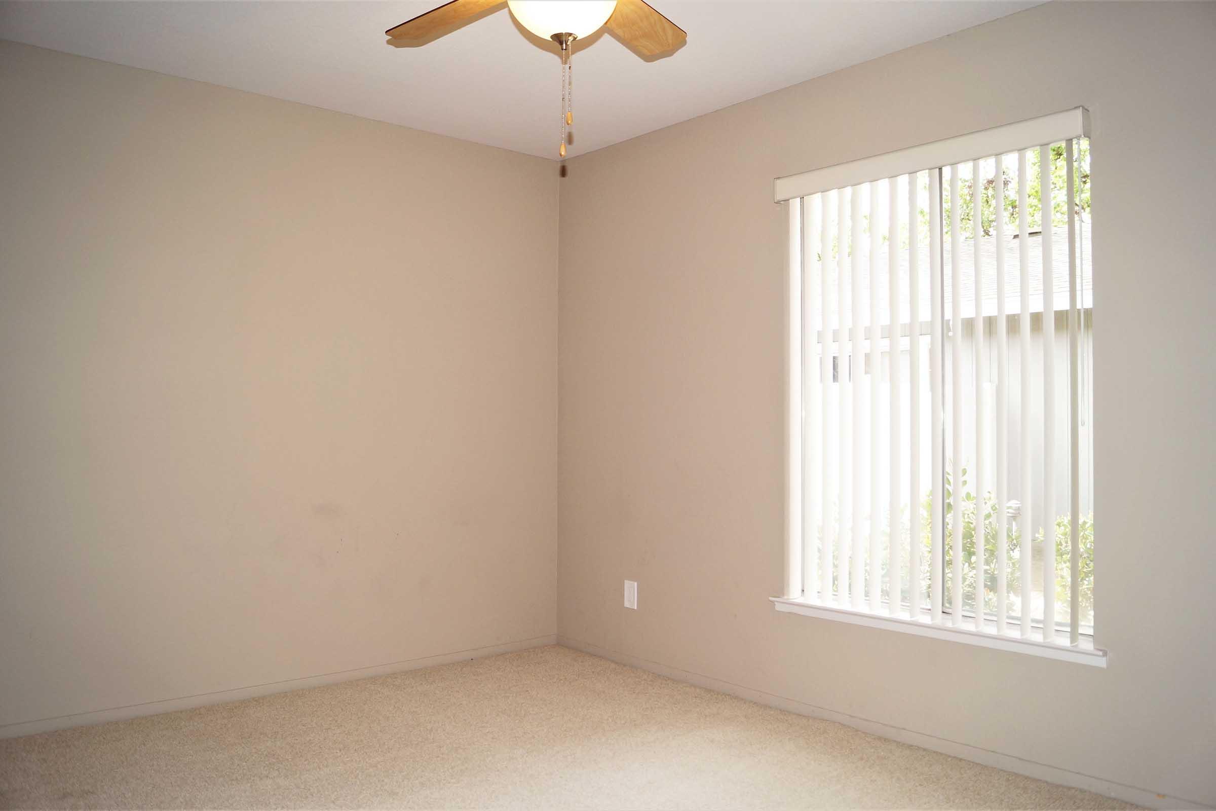 A vacant room with light beige walls and a ceiling fan. There is a window with vertical blinds that allows natural light to enter, illuminating the beige carpeted floor. The room appears simple and uncluttered, suitable for various purposes.