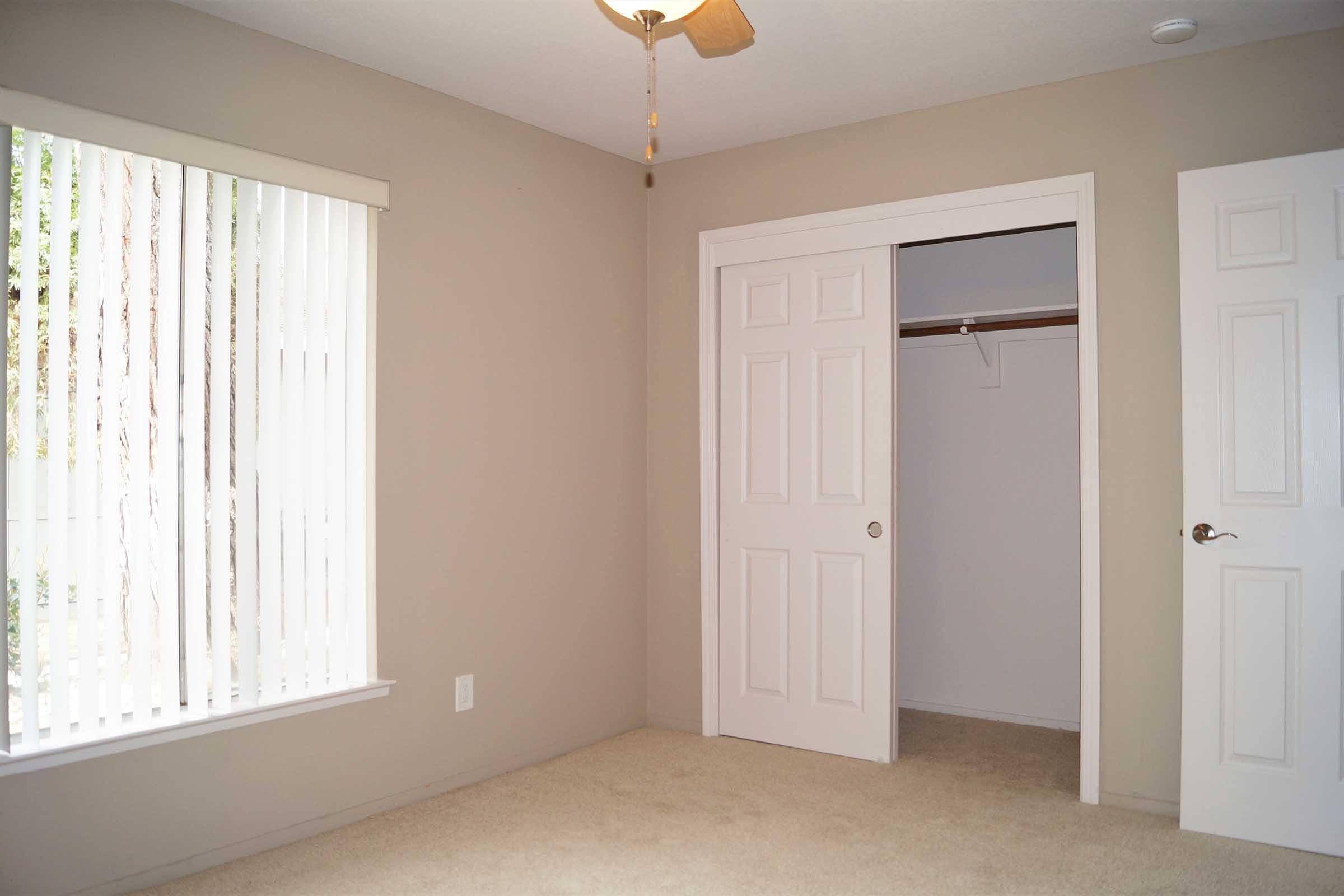 Empty room with beige walls and carpet. A window with vertical blinds allows natural light to enter. There is a ceiling fan and a closet with a white door slightly open. The overall atmosphere is clean and simple, suitable for a bedroom or office space.