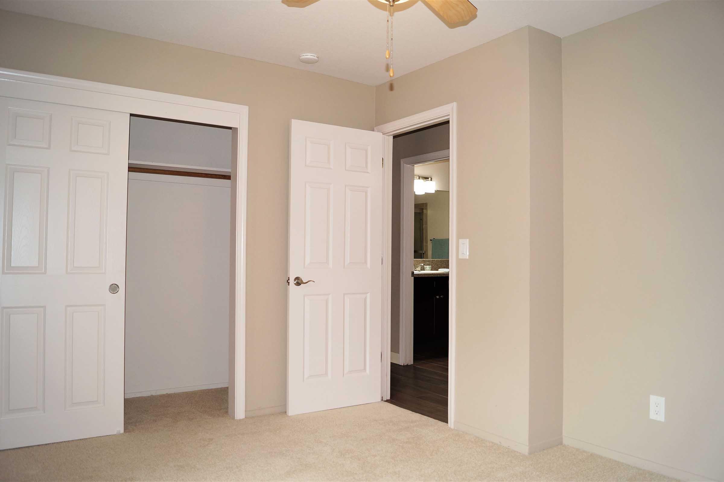 A view of a bedroom featuring light beige walls and carpeting. There are two closet doors on the left, a doorway leading to another room on the right, and a ceiling fan overhead. The space is bright and uncluttered, creating an open and inviting atmosphere.