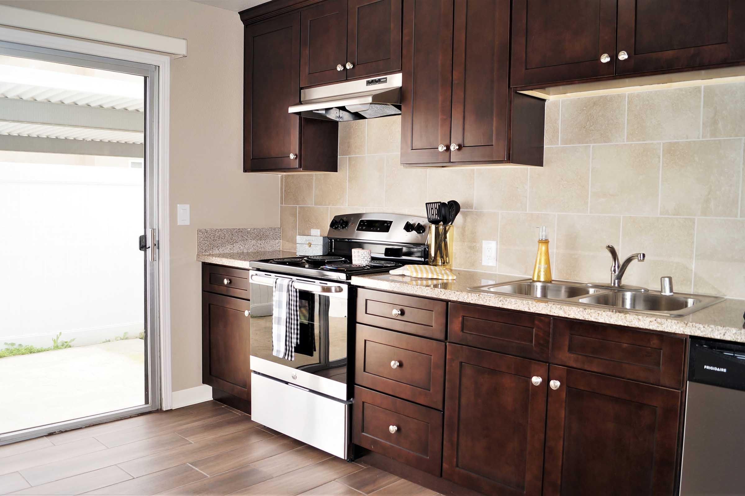 A modern kitchen featuring dark wood cabinetry, a stainless steel stove, and a double sink. The countertop is made of light granite. There is a sliding glass door leading to an outdoor area, creating an open and airy atmosphere. The kitchen is well-lit, with beige tiles on the walls and a clean, organized appearance.