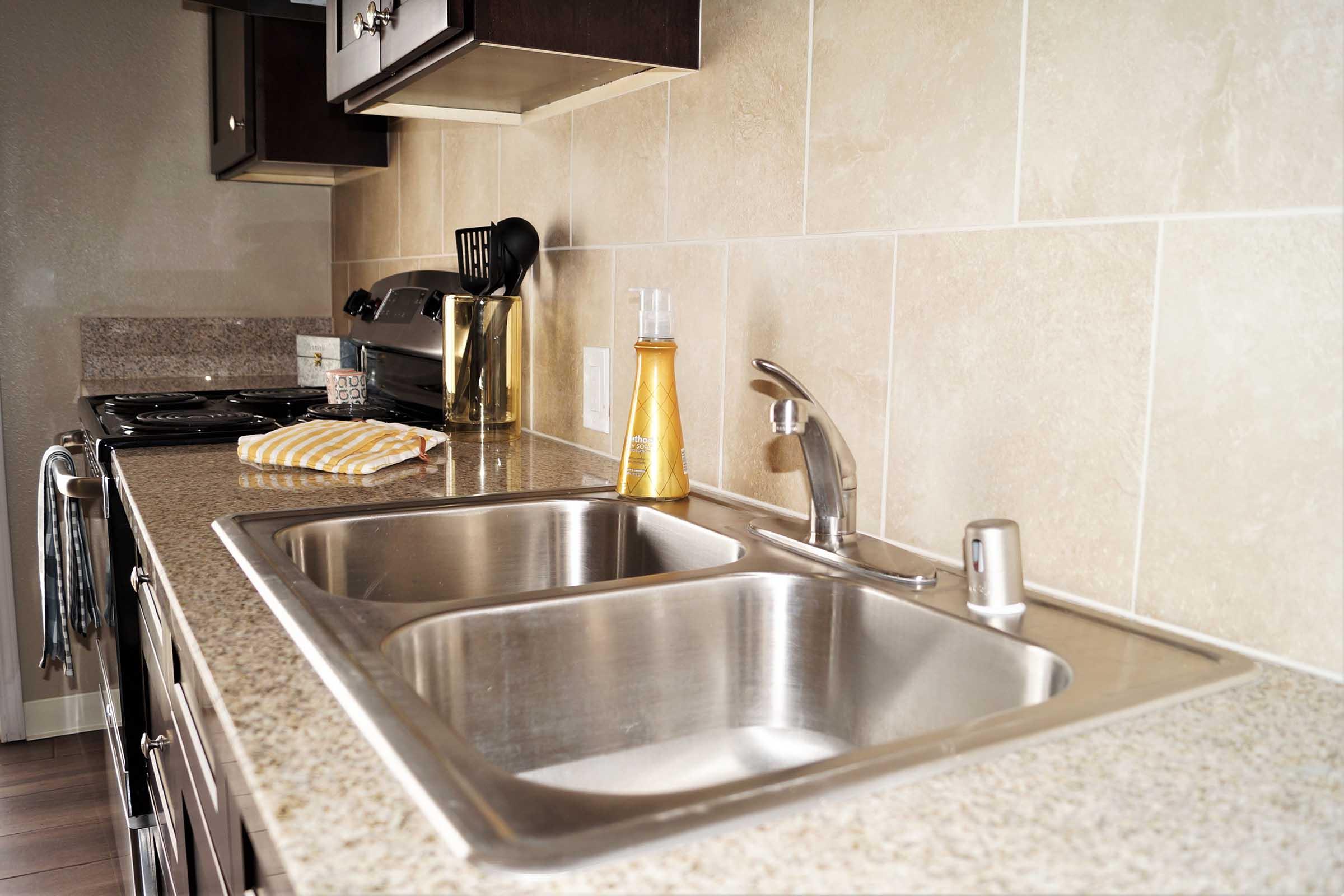 A modern kitchen sink area featuring two stainless steel basins, a sleek faucet, and a countertop with a light brown granite finish. A dish towel is placed on the counter, next to a soap dispenser. The kitchen has dark cabinetry and an open layout.