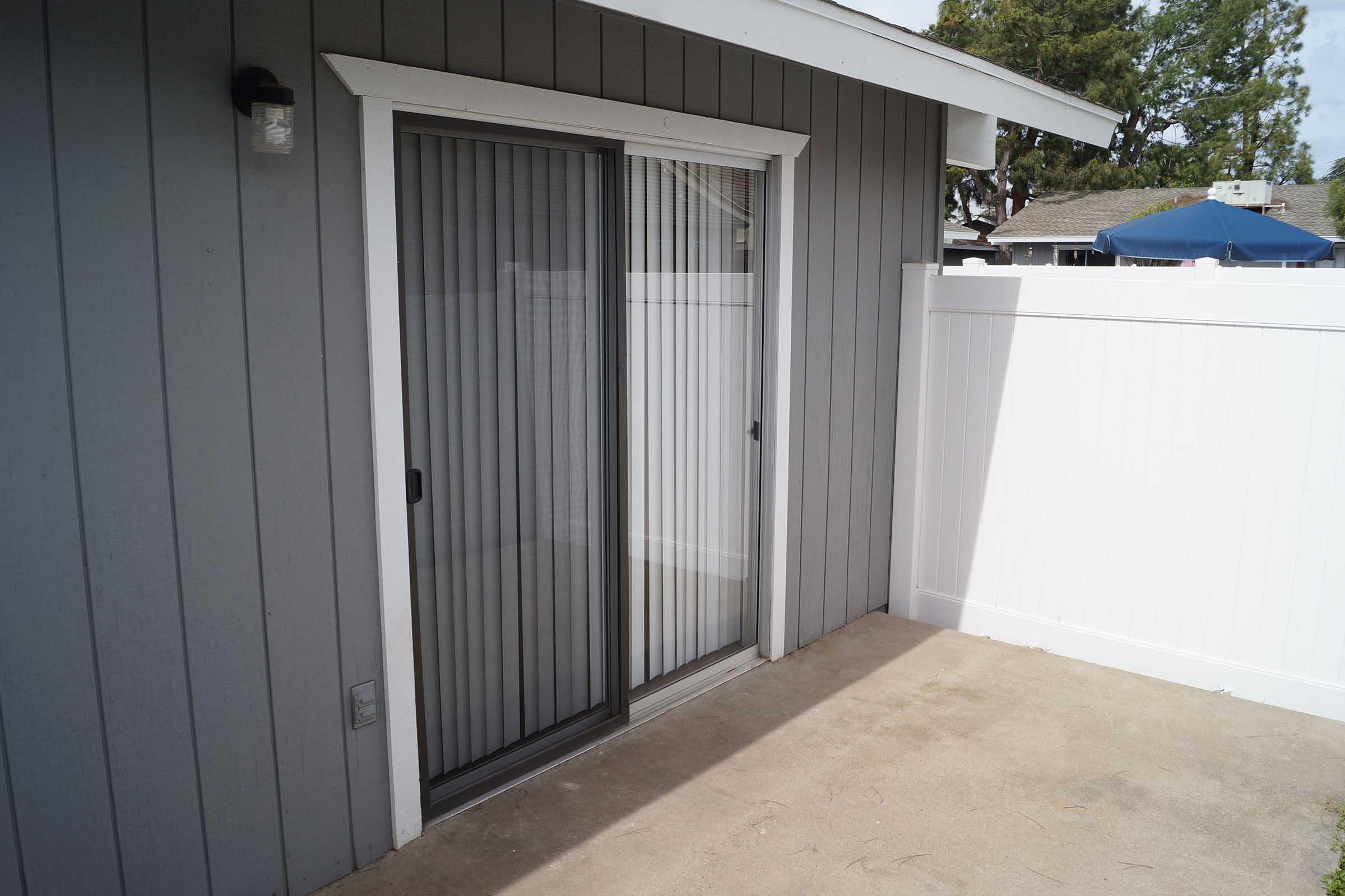 Sliding glass door with vertical blinds in a gray exterior wall, leading to a small outdoor area. There is a white fence in the background, with a patio visible. The space is well-lit and features a simple, clean design.