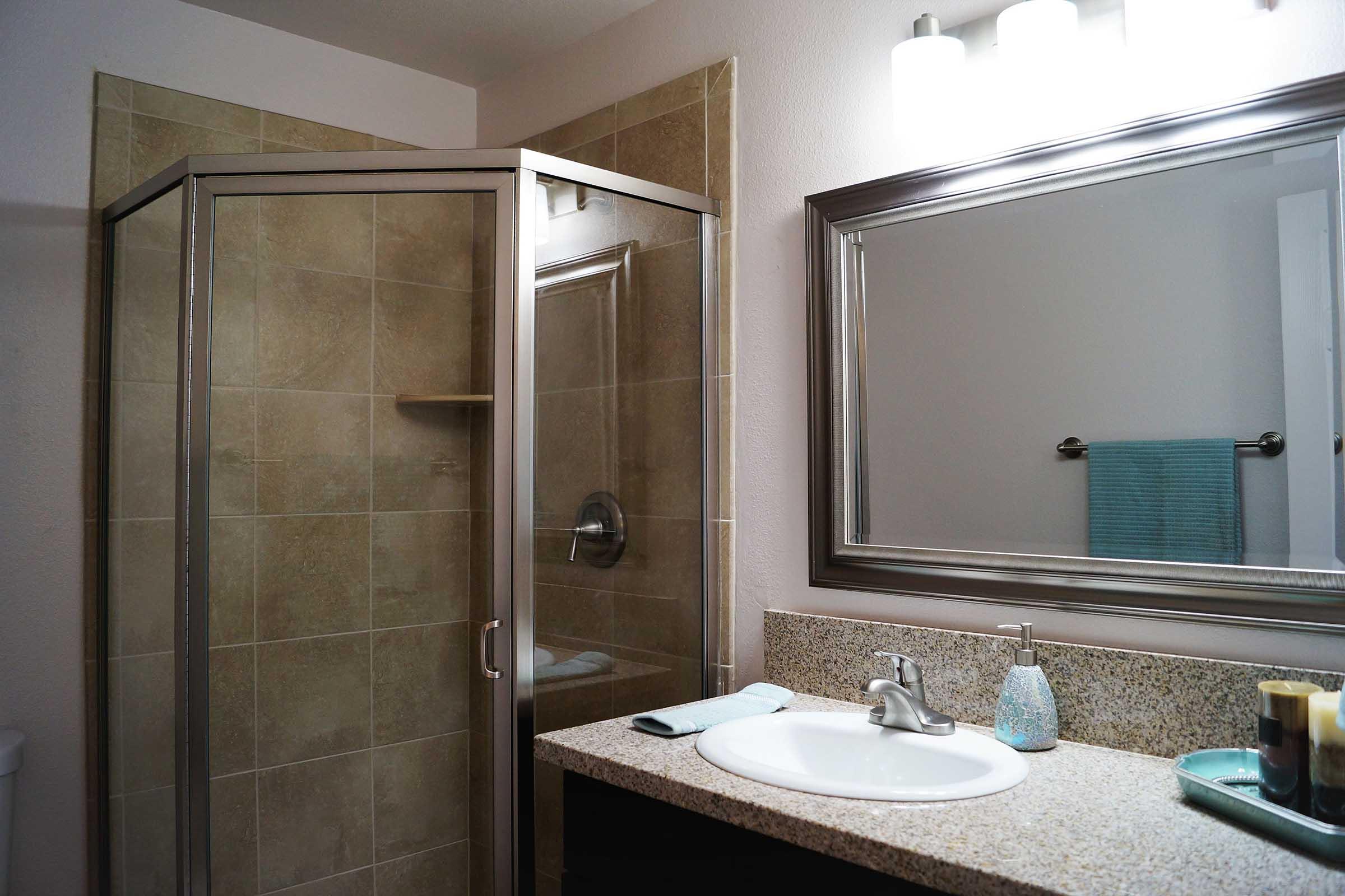A modern bathroom featuring a glass shower enclosure, a beige tiled wall, a granite countertop with a sink, a mirror with a silver frame, a light fixture above, and a few decorative items. A light blue towel hangs on a rail, and a toilet can be seen in the background.