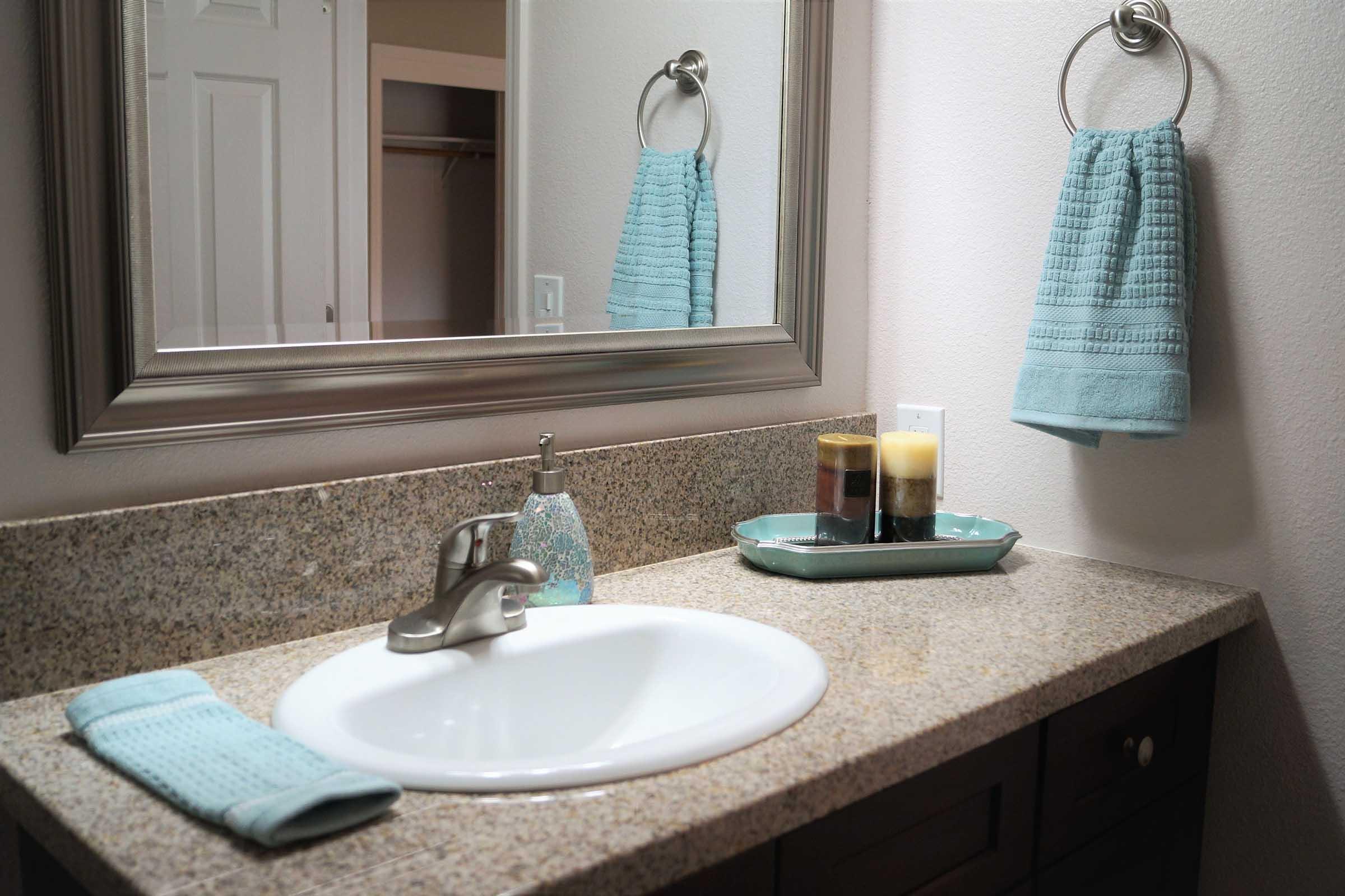 A modern bathroom sink with a light granite countertop, featuring a faucet, a soap dispenser, and a tray holding two candles. Nearby, a folded towel in a soft blue hue hangs on a towel ring, and another matching towel is neatly placed on the countertop. A large mirror reflects the space.