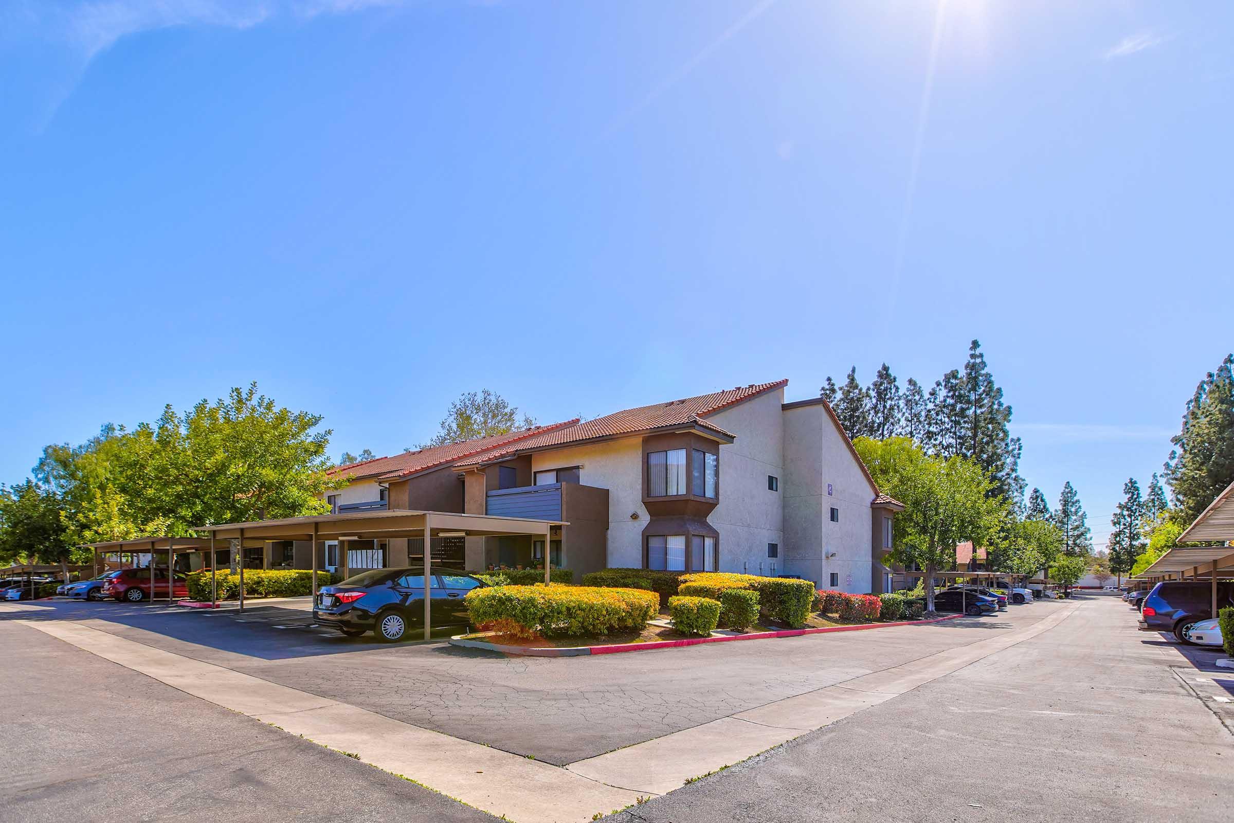 A view of a residential apartment complex with two-story buildings. The foreground features a parking lot with several cars and covered parking spaces. Lush green shrubs and trees surround the complex, under a clear blue sky with sunlight. The scene conveys a peaceful suburban environment.