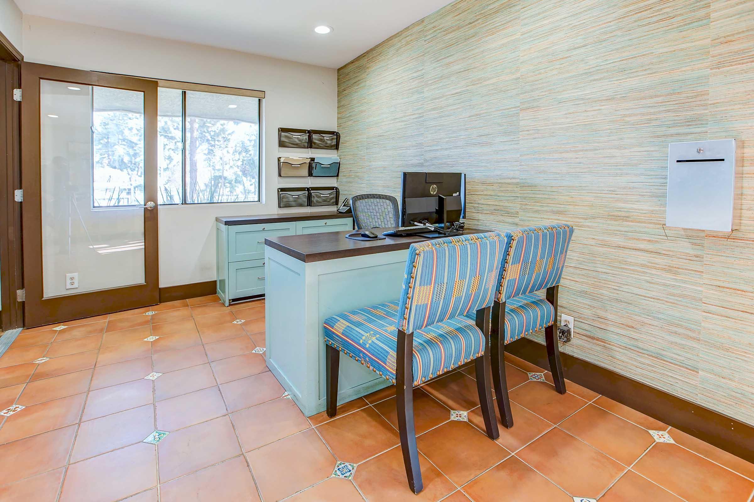 A modern office space featuring a desk and two patterned chairs. The room has light brown tiled flooring and natural light coming from a window. There is a computer on the desk and a wall-mounted mailbox nearby. The walls are decorated with a textured light-colored material.
