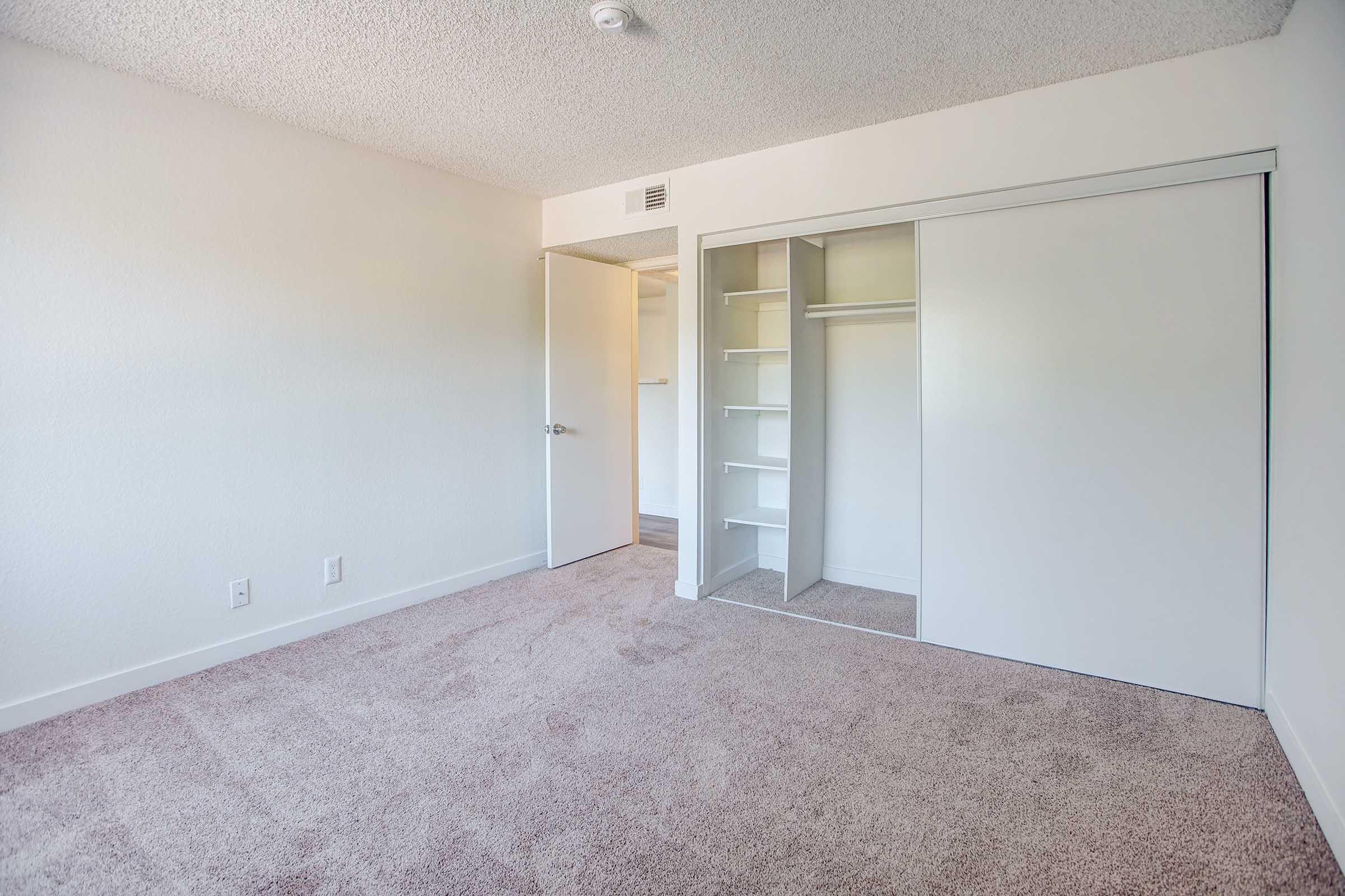 A simple, empty bedroom with light-colored walls and carpet. It features a closet with sliding mirrored doors and built-in shelves. A door leads to another area, and the overall space appears bright and airy, with natural light illuminating the room.