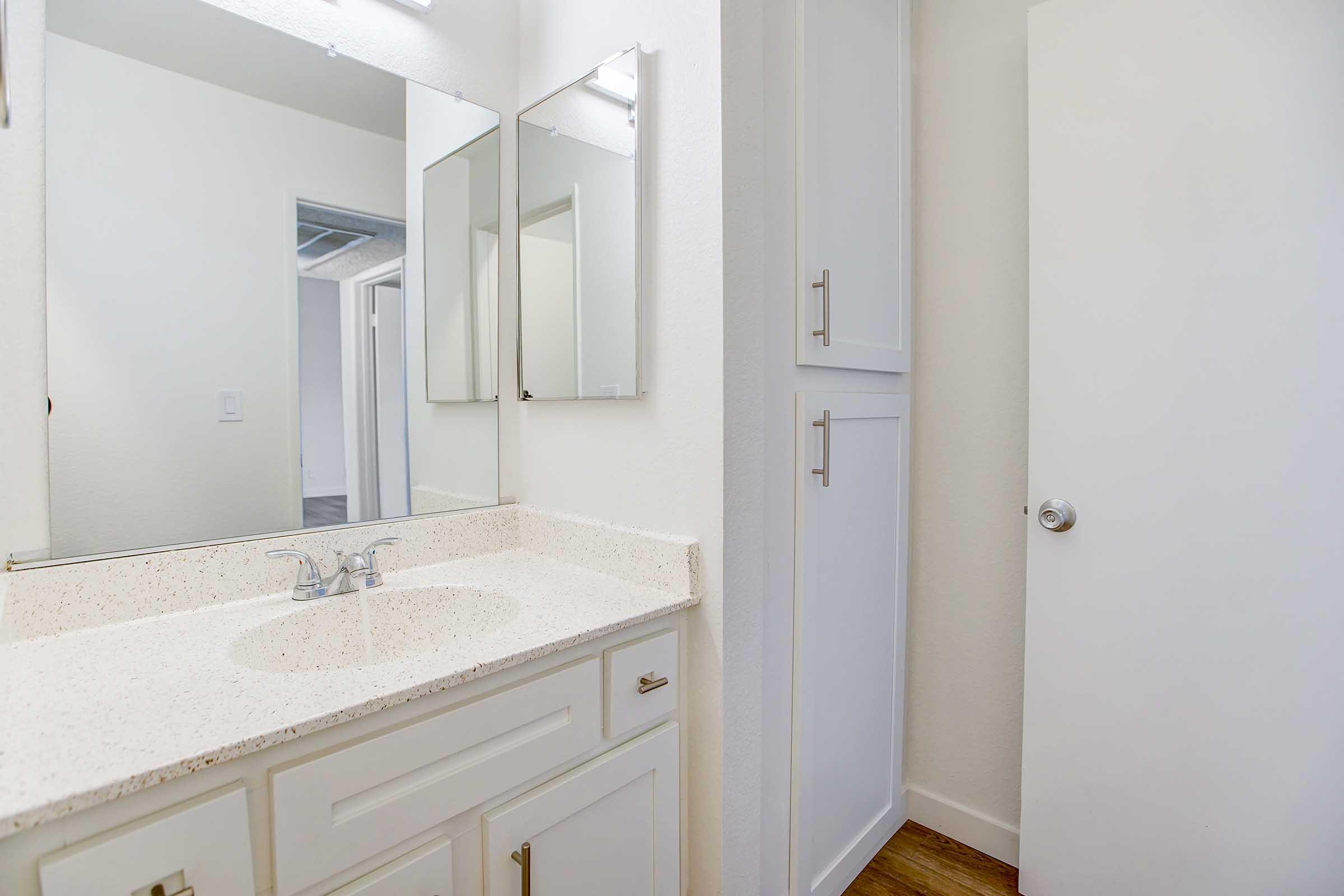 A well-lit bathroom featuring a white countertop sink, two mirrors above, and a cabinet. The walls are painted light, and there is a closed door on the right. The flooring appears to be wood-like. The space is clean and modern, with a minimalist design.