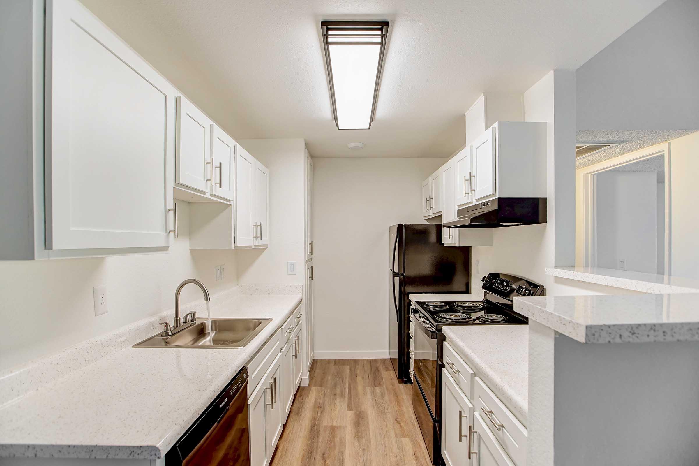 A modern kitchen featuring white cabinetry, a stainless steel sink, and a black refrigerator. The space includes an oven and stovetop, with light-colored countertops and a ceiling light fixture. The flooring is a light wood laminate, creating a bright and inviting atmosphere.