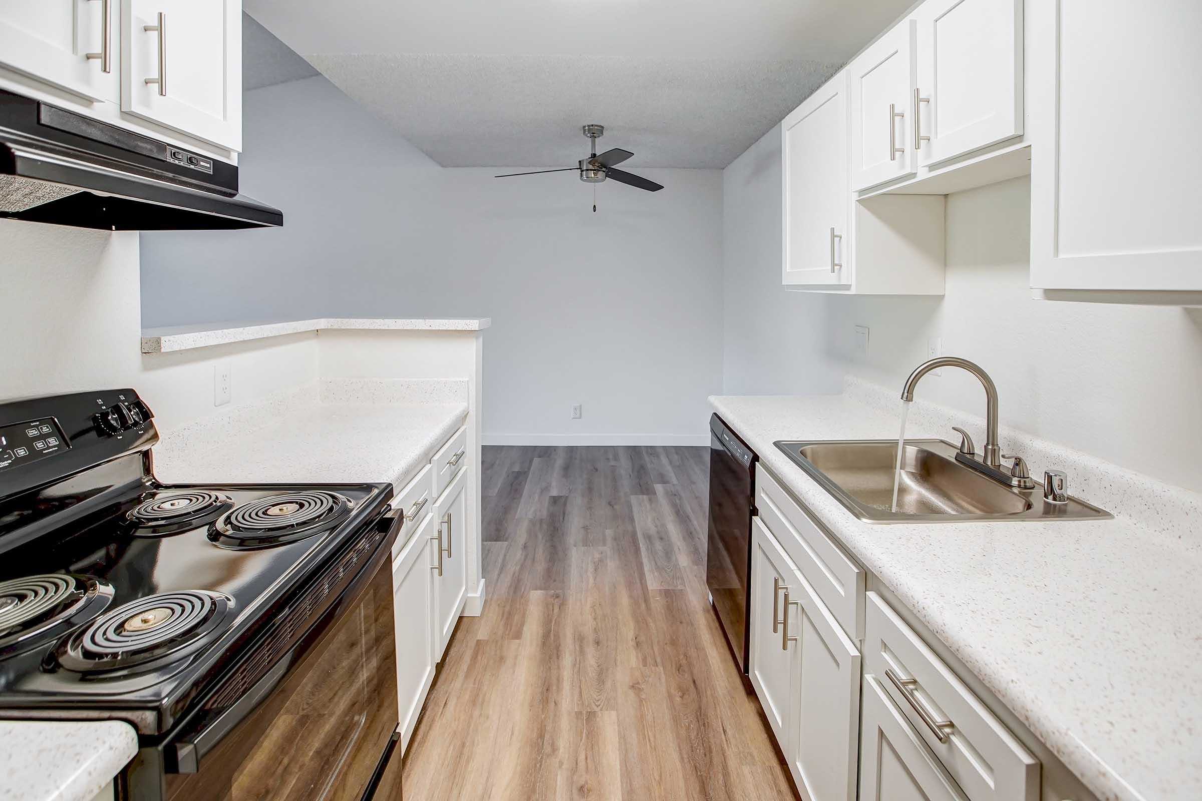 Modern kitchen featuring white cabinets, black appliances including a stove and dishwasher, and a stainless steel sink. The flooring is light wood laminate, and there is a ceiling fan in the background. The space is well-lit and open, leading into a living area with a clean, minimalist design.